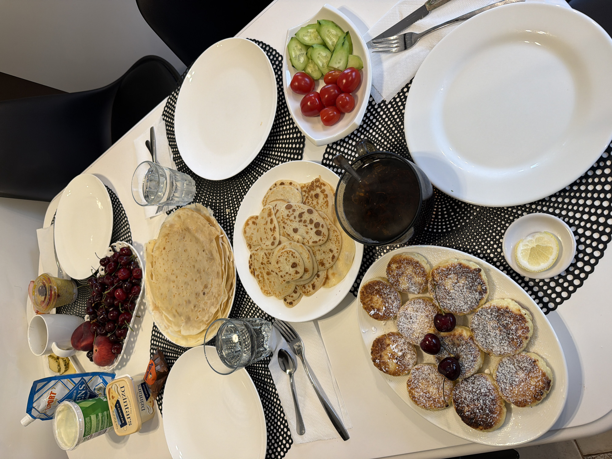 The image shows a breakfast table set for a meal. There are plates, cutlery, glasses of water, and various food items. The food includes a stack of large crepes, a plate of smaller pancakes dusted with powdered sugar and garnished with cherries, a bowl of cherries and a peach, a dish of cherry tomatoes and cucumber slices, a tub of yogurt, a package of butter, a small cup of mustard, and a cup of tea or coffee. The table is set on a white surface with black and white placemats. Two black chairs are visible in the background. The scene appears to be indoors, possibly in a dining area. No people are visible in the image. The location is identified as Baloži, Latvia.