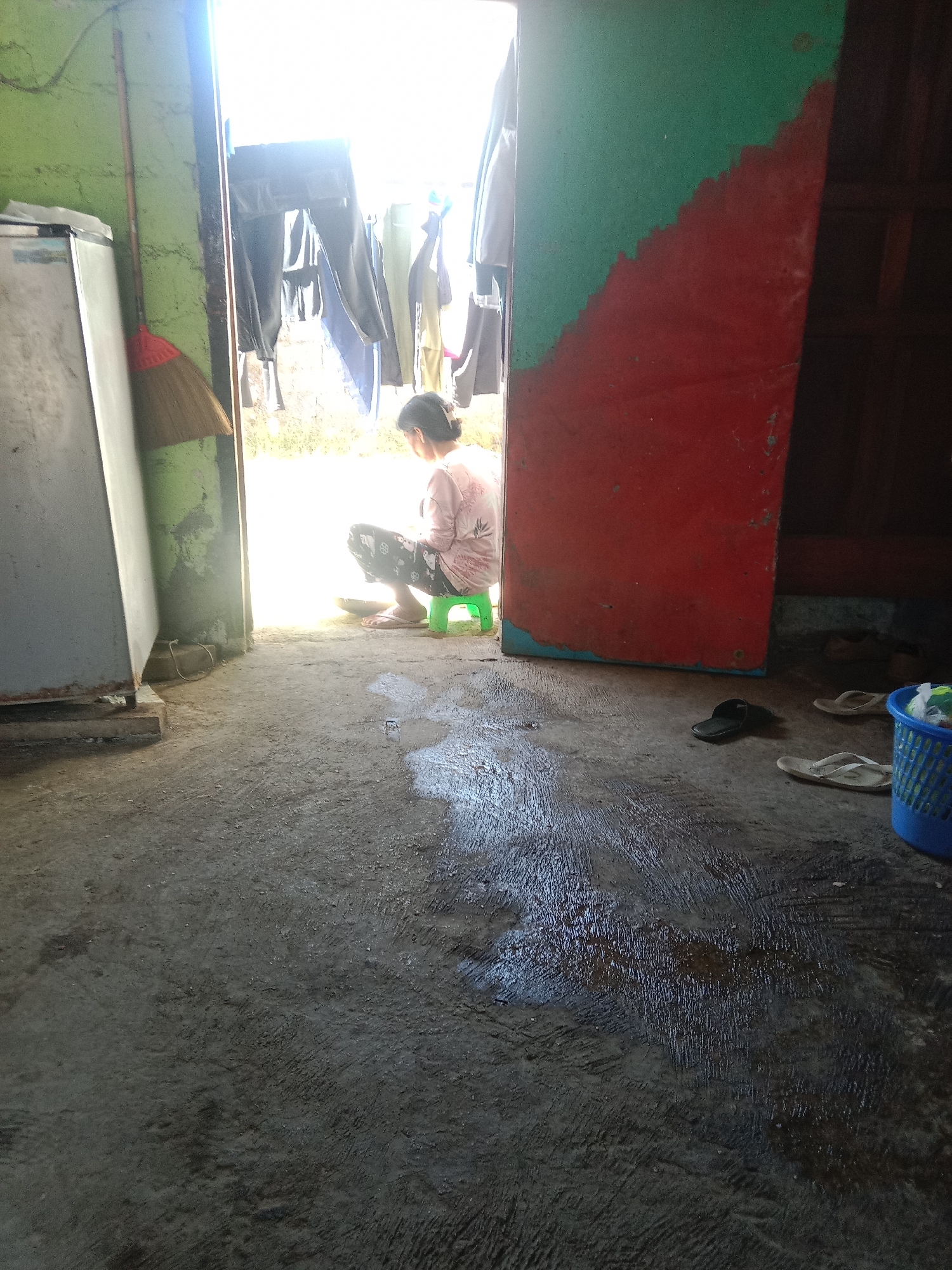 The image depicts an interior scene, possibly a doorway or opening into an outdoor area. In the foreground, a concrete floor is wet and reflective, showing patches of dark liquid. To the left, a metallic appliance, likely a refrigerator, stands against a green-painted wall. A broom with a red head leans against the wall next to it.
In the middle ground, a person is seen crouching on a small, bright green stool, facing away from the camera. They are wearing a light-colored patterned shirt and dark pants. The person appears to be holding a bowl or shallow container.
The background is dominated by bright light, suggesting an outdoor space or an area with strong illumination. Clothes are hanging on a line in the background, some of which are dark-colored pants. To the right, a large red-painted surface with a diagonal green section forms a wall or partition. Further to the right, a blue plastic basket filled with items is visible. Several pairs of flip-flops and a single black sandal are scattered on the floor near the red partition.
This scene is located in Leksono, Indonesia.