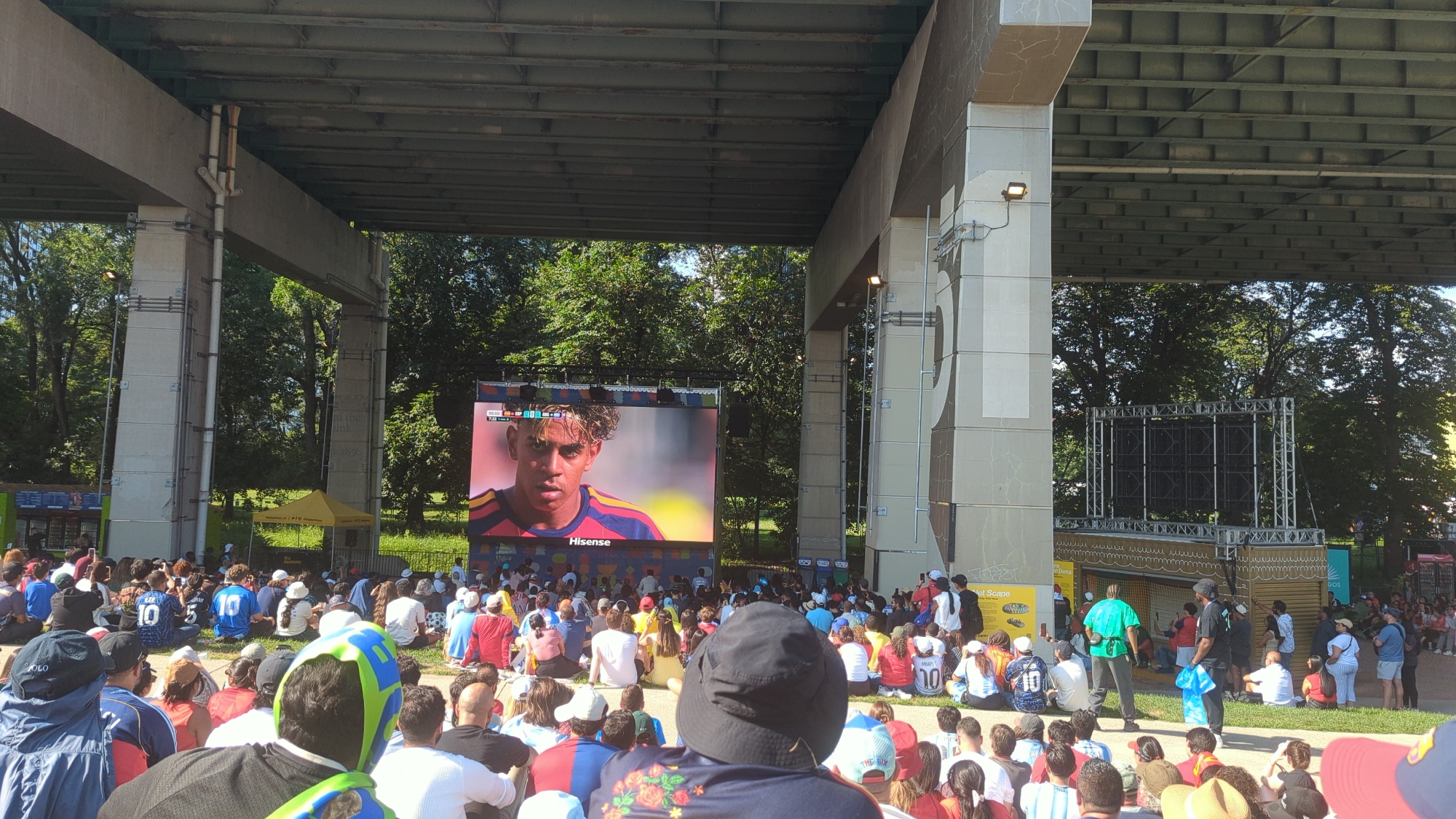 The image depicts an outdoor public viewing of a soccer match, likely during a summer day, in an urban setting. A large screen, branded "Hisense," displays a close-up of a young man with curly hair wearing a soccer jersey. Below the screen, a crowd of people, many wearing soccer jerseys of various colors including blue, red, and white, are seated on the ground. The setting appears to be beneath a large overpass, with concrete pillars and a metal structure overhead. Trees and foliage are visible in the background, suggesting a park-like or green space within the city. A yellow tent with signage is also present, possibly indicating vendors or event information. The overall atmosphere suggests a communal gathering to watch a sporting event. This scene is likely taking place at the Stackt Market in Toronto, Canada, which is known for hosting outdoor events and screenings under an overpass.