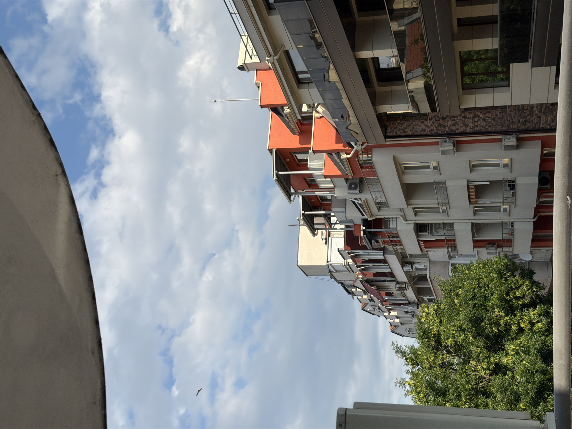 The image depicts a side view of a modern apartment building with balconies and a few windows visible. The building has sections painted in white and vibrant orange, with red accents. The balconies have metal railings and some have awnings extended. There are several air conditioning units mounted on the exterior walls. In the foreground, a lush green tree with yellowing leaves is partially visible on the left. The background features a blue sky with scattered white clouds. A single bird is captured flying in the sky. The building appears to be located in an urban environment, likely a residential area in Plovdiv, Bulgaria, given the architectural style. The overall scene suggests a daytime view, possibly in late summer or early autumn.