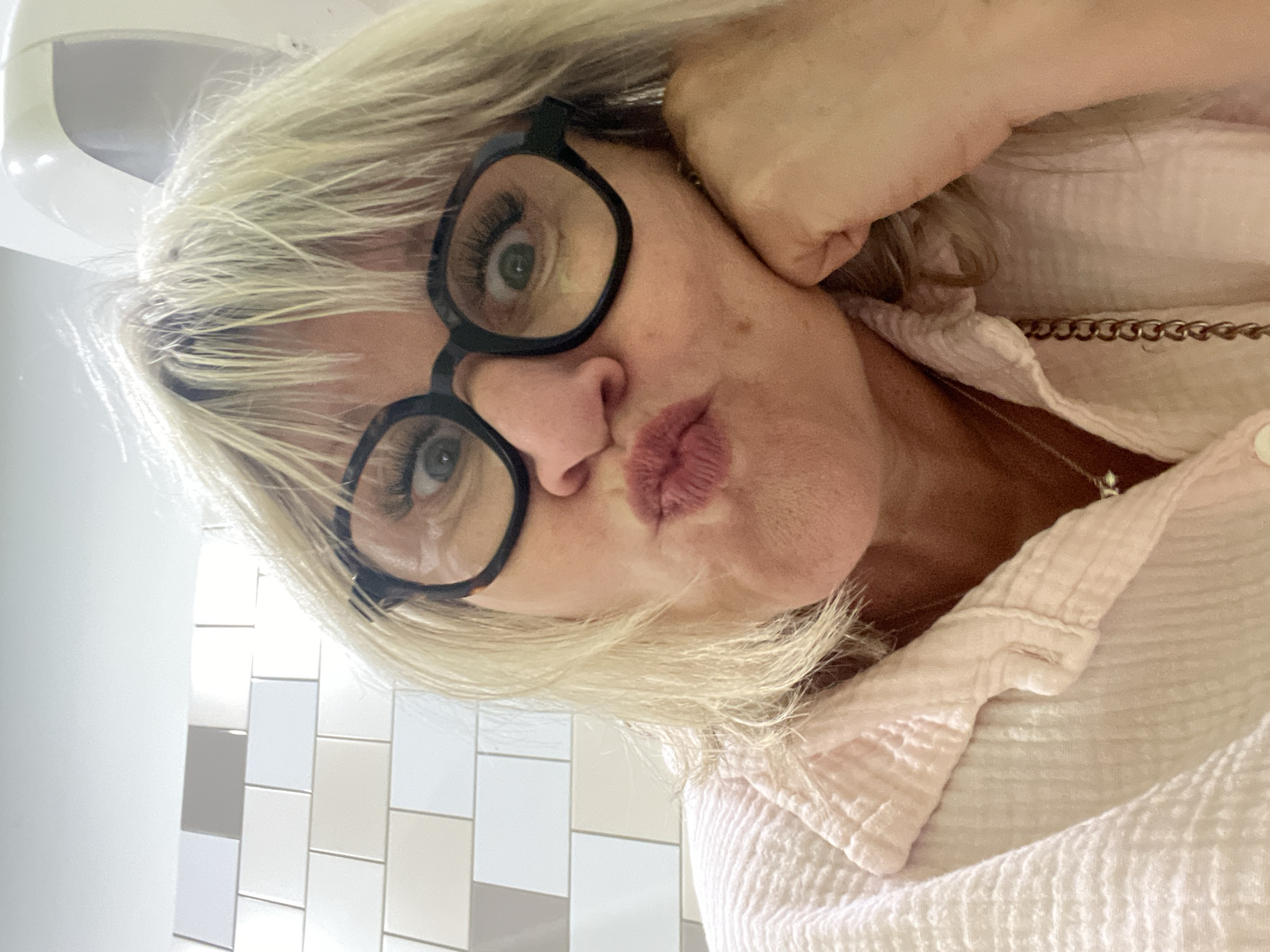 A selfie was taken in a room with tiled walls in Rousset, France. An adult woman with light blond hair is posing with puckered lips and black glasses. She is wearing a light pink blouse and a gold chain necklace. Her left hand supports her head. A part of a white object is visible on the left side of the frame. The tiles on the wall are white, gray, and beige.
