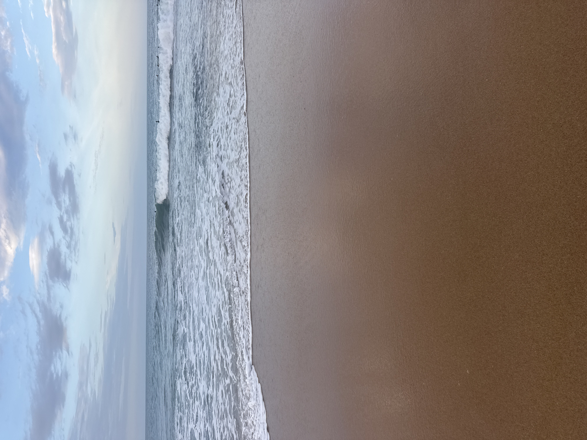 The image depicts a serene beach scene at Arugam Bay, Sri Lanka. The foreground is dominated by the wet, brown sand of the beach, reflecting the soft light of the sky. In the mid-ground, gentle waves with white foam are breaking and receding onto the shore, creating a textured line where the water meets the sand. The ocean stretches out into the background, appearing a calm blue-green. Further out on the water, several surfers are visible, scattered across the expanse, either waiting for waves or riding them. The horizon is defined by the meeting of the ocean and the sky. The sky itself is a blend of pale blue and white, with scattered, wispy clouds that suggest either early morning or late afternoon light. The overall atmosphere is tranquil and natural, characteristic of a coastal landscape.