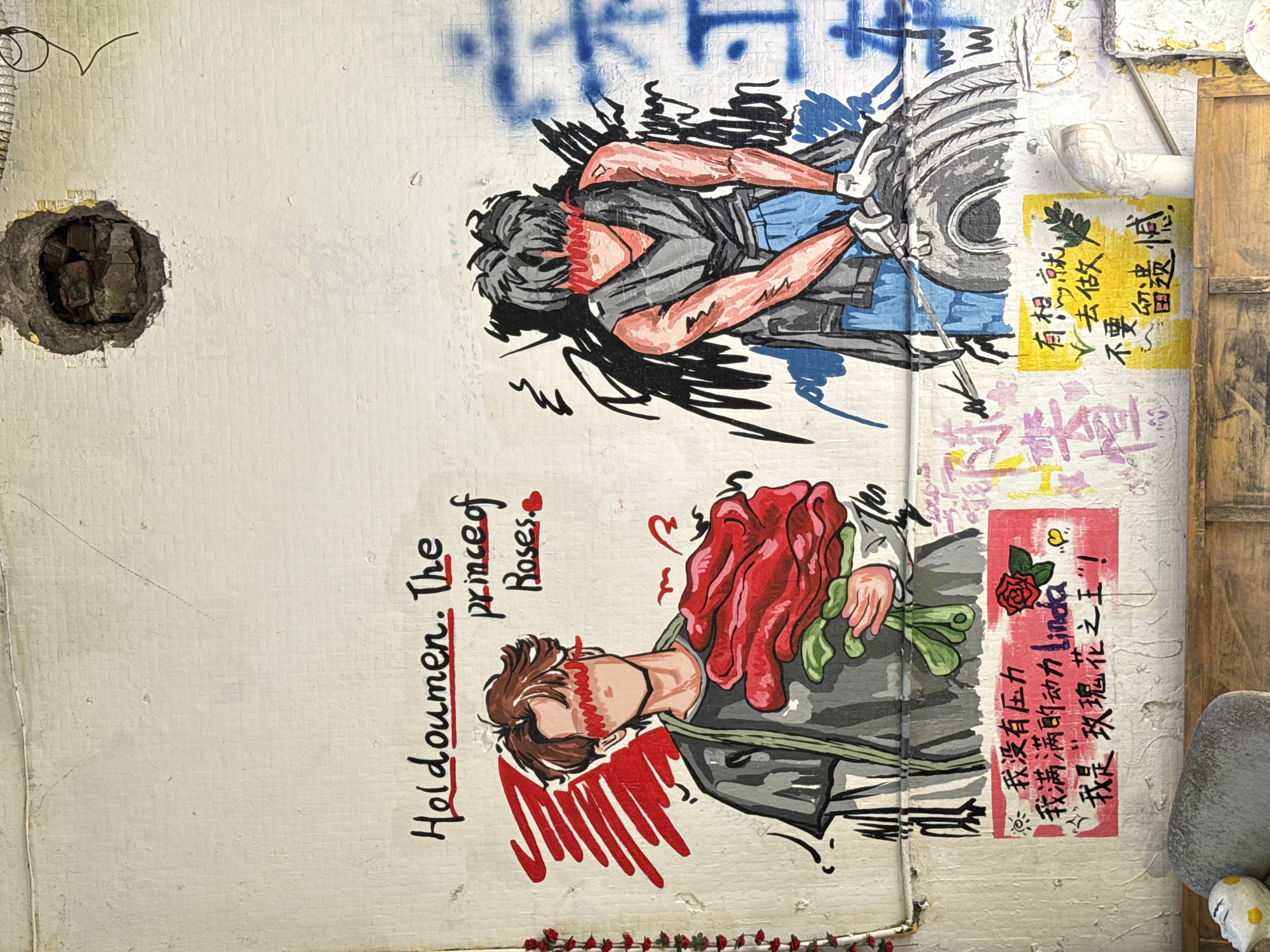 The image is of a wall in Jiulongpo District, China. The wall has a hole in it on the left side. The rest of the wall is covered in graffiti. There are two images, one above the other. The top image is of a person lying down, with their arms reaching up to touch a tire. The person is wearing a gray shirt and blue pants. The bottom image is of a person holding a bouquet of red roses. The person is wearing a gray shirt and black pants. There are also some Chinese characters written on the wall. The text is in black and red ink. There are also some stickers on the wall. There is also a wooden door frame to the right.