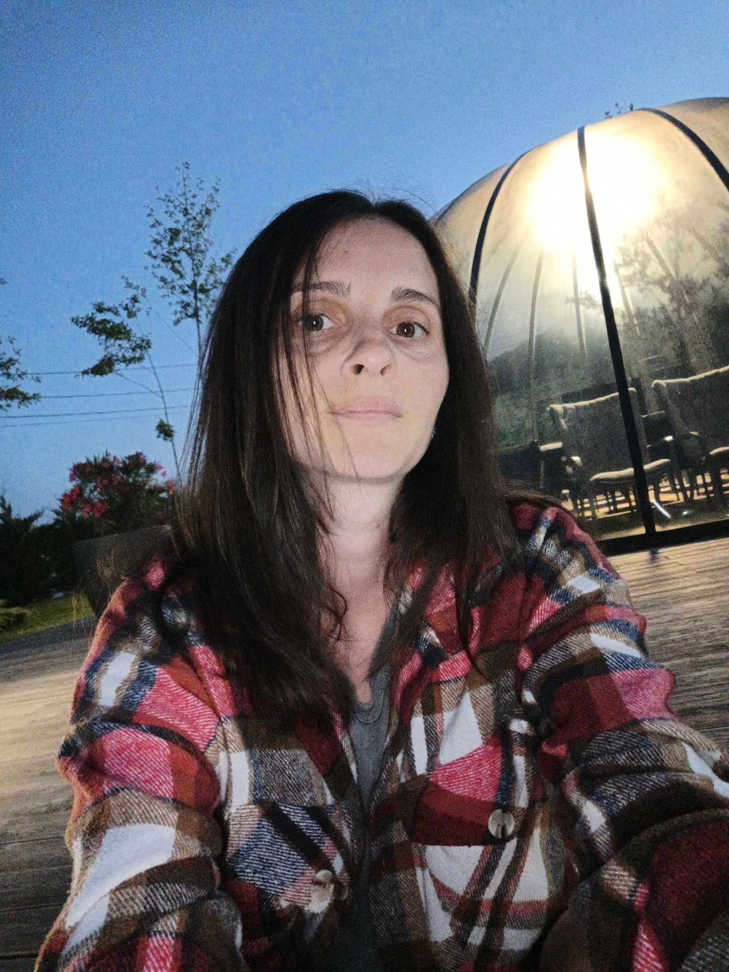 A woman is taking a selfie outdoors, presumably in Pshada, Russia, as indicated by the prompt. The sky is a clear blue, suggesting it is either early morning or late evening. In the background, a large, transparent geodesic dome glows from within, casting a warm light. Inside the dome, several chairs are visible. The woman is wearing a red, brown, and white plaid shirt over a grey shirt. She has dark brown hair that falls around her shoulders. The setting appears to be on a wooden deck or patio.