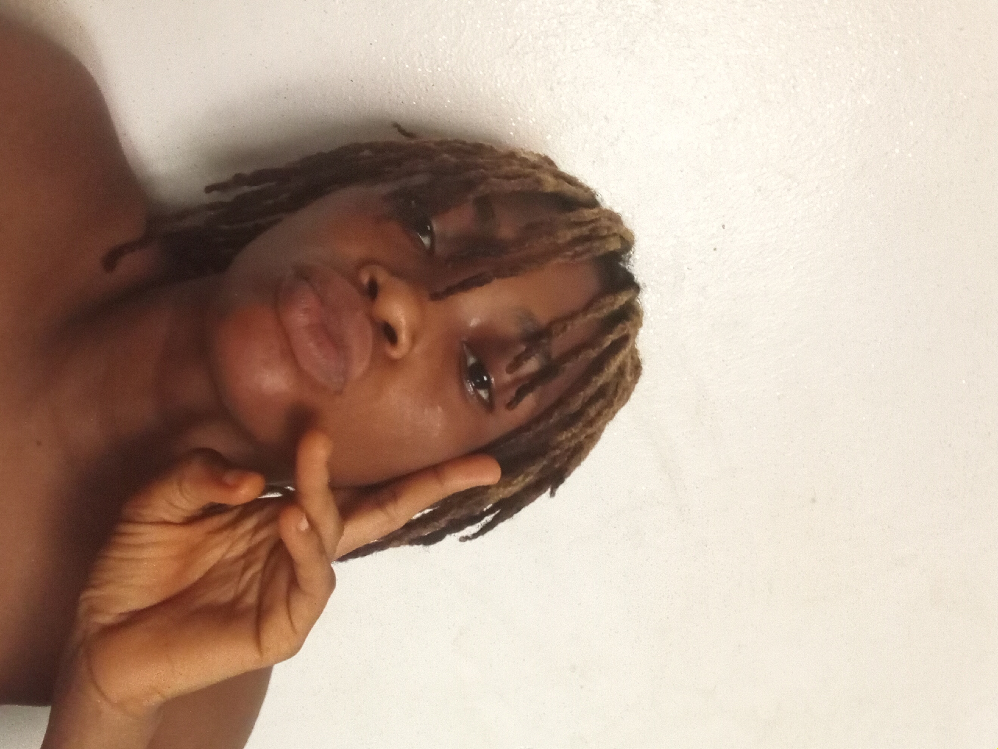 The image depicts a dark-skinned individual with dreadlocks, pursing their lips, and making a peace sign with their hand. The background is a plain white surface. The location and city are unknown.