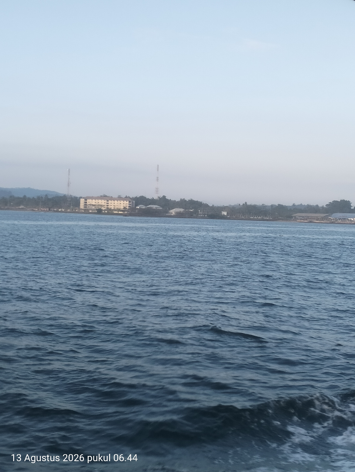 The image depicts a view of the sea with the city of Kendari, Indonesia visible in the distance. The sky is clear and pale blue, suggesting it is early morning or late afternoon. The water is a deep blue with gentle ripples. In the background, several buildings are visible, including a large, multi-story hotel or apartment complex, along with what appear to be telecommunication towers. Trees line the coast, and a hilly landscape can be seen further inland. The timestamp on the image indicates it was taken on August 13, 2026, at 06:44. There are no people visible in the image.