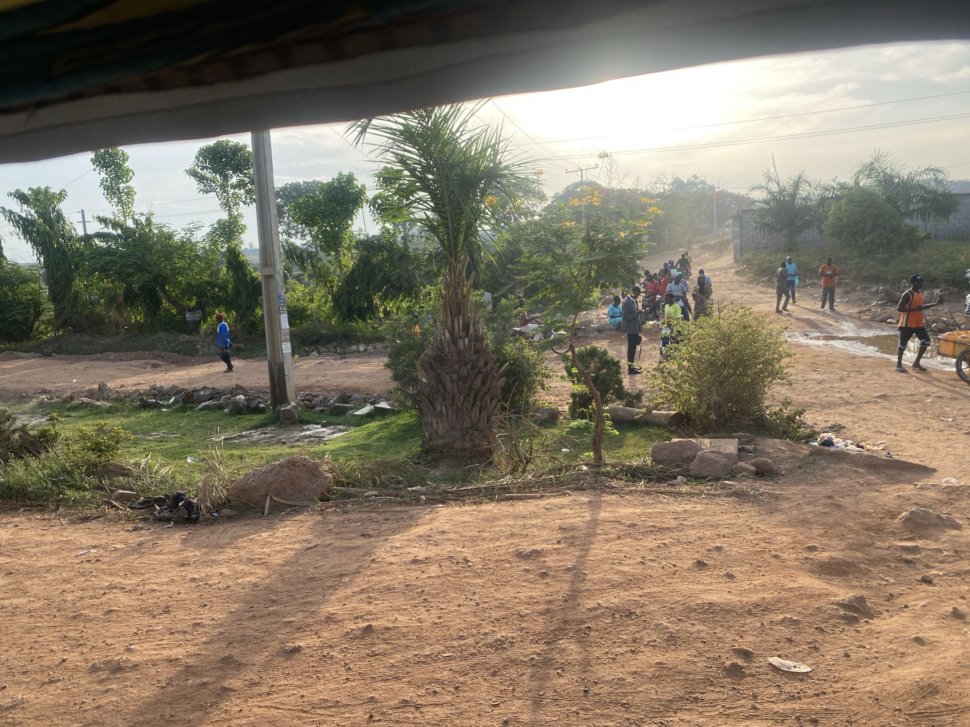 The image depicts a dirt road intersection in Kuchingoro, Nigeria. The foreground is dominated by reddish-brown dirt. Vegetation, including a palm tree and other green foliage, is present. A group of people is visible further down the road. Some individuals are carrying yellow containers, possibly water. A utility pole stands on the left side of the road. A concrete wall partially surrounds the dirt road, showing a possible border for land.