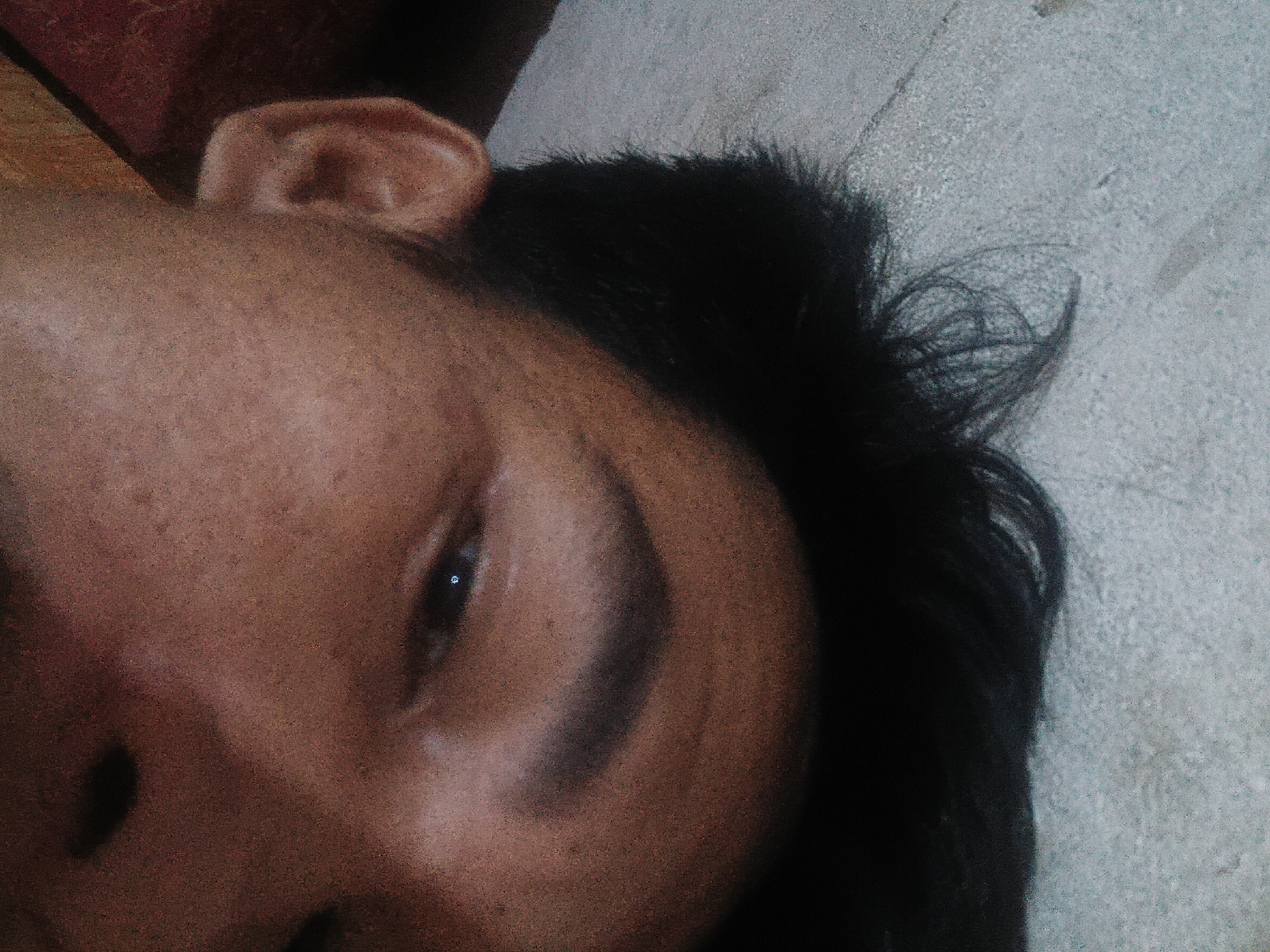 The image is an extreme close-up, angled shot of a person's face, lying on their side. The subject's right ear is visible in the upper left quadrant of the image, and their right eye, with a visible pupil reflecting light, is in the center. Dark hair frames the face. The background appears to be a textured surface, possibly a pillow or blanket, with a reddish-brown object in the upper left corner. The lighting is dim and appears to be coming from above and slightly to the left, casting a shadow across the face. The image quality is grainy and of low resolution. There is no discernible information in the image to indicate the location of Prabumulih, Indonesia.