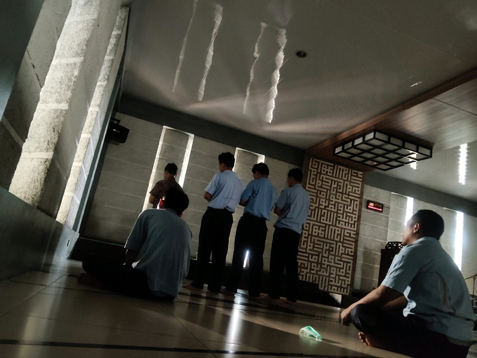 In the city of Mekarmukti, Indonesia, this image depicts several young males praying inside a mosque. The individuals are dressed in light blue shirts and dark pants. One individual is seated on the floor in the foreground, facing away from the camera. Behind him, four other males stand in a line, facing the same direction. The mosque features a tiled floor and walls with decorative Islamic calligraphy. Sunlight streams through tall windows, casting distinctive patterns of light and shadow across the room and the ceiling. A modern light fixture is mounted on the ceiling. There is a digital clock mounted on the wall. A discarded face mask lies on the floor in the foreground.