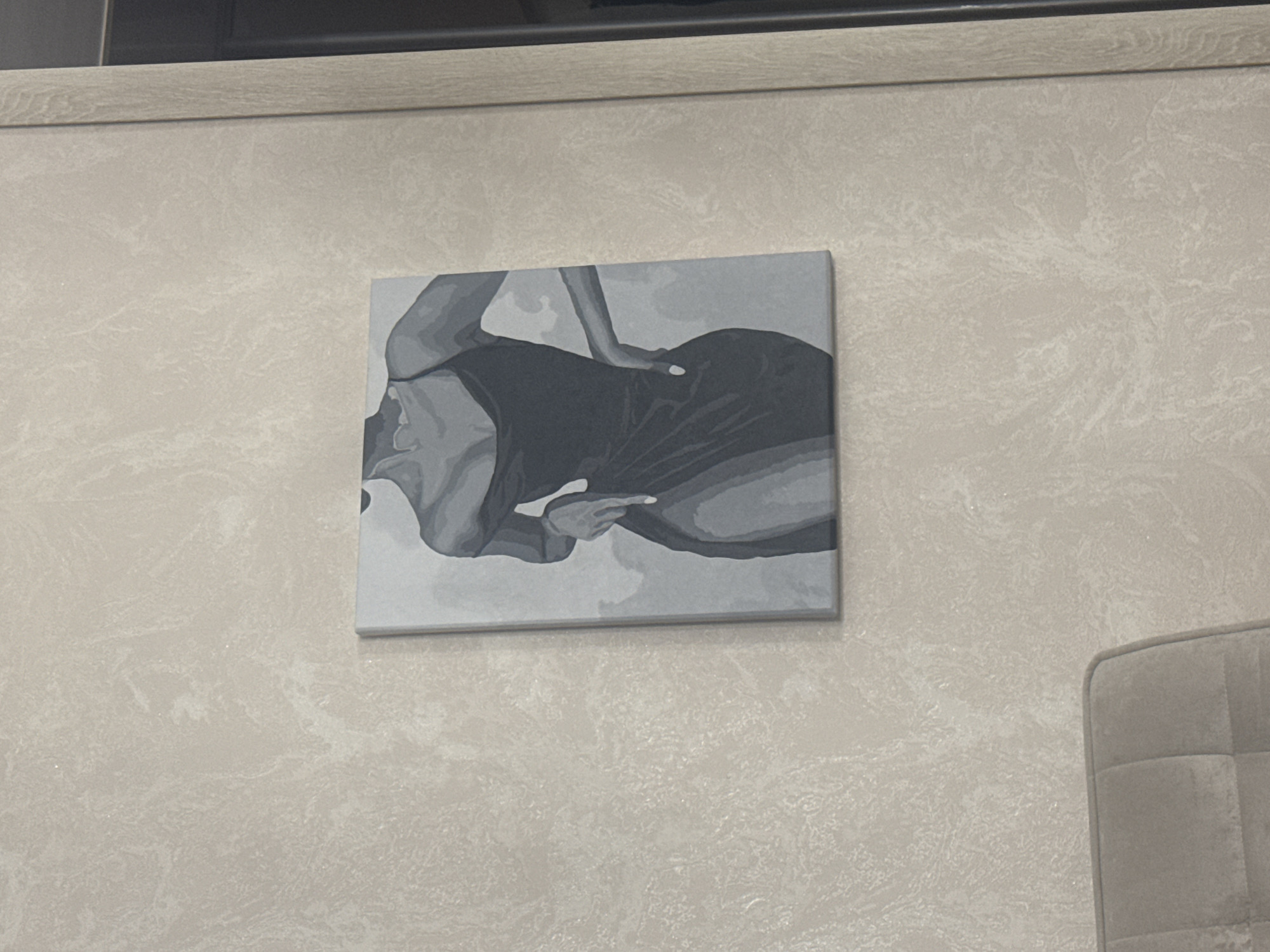 A grayscale painting of a woman in a dress is hanging on a textured wall. The woman is depicted from the chest down, with her left hand on her hip and her right hand touching her thigh. She is wearing a slip dress with thin straps, and the dress has a thigh-high slit. The painting is rectangular and appears to be on canvas. The wall is a light beige color with a subtle, swirling pattern. To the right of the painting, a sliver of a wooden door frame is visible. In the lower left corner of the image, the top of a light brown upholstered chair with tufted detailing can be seen. The overall scene suggests an indoor residential setting. The location is not specified in the image or its crops.
