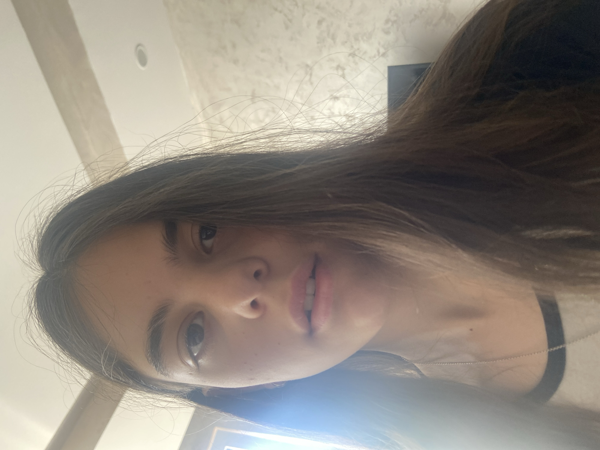 A selfie of a young woman with long brown hair. Her skin appears pale and her eyes are brown. She is wearing a black and white shirt and a thin necklace. The background shows a textured wall and a ceiling with a recessed light fixture. The setting seems to be indoors, potentially inside a home.
There is no indication of location in the city of Dragovishtitsa, Bulgaria.