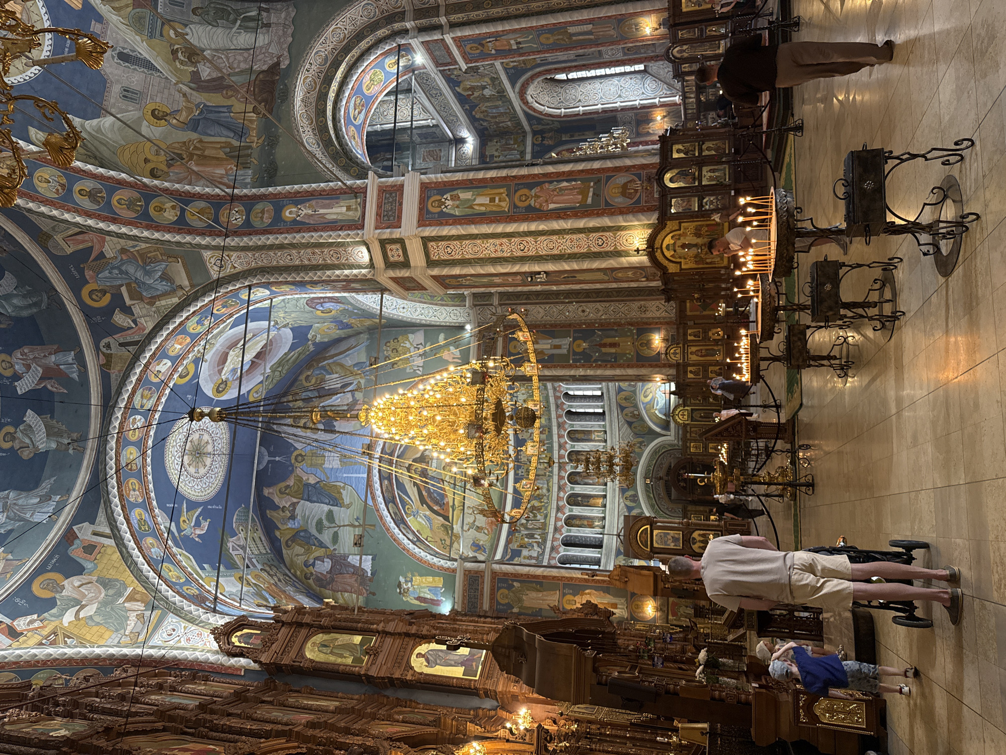 The image captures the interior of Alexander Nevsky Cathedral in Nizhny Novgorod, Russia. Ornate religious paintings cover the vaulted ceilings and arched walls. A large, gilded chandelier hangs centrally. In the foreground, wooden structures, adorned with icons, line the lower walls. Several people are visible: a man in a beige shirt and khaki shorts stands near a stroller, and a child in a blue shirt is nearby. Candles are lit on stands, and other individuals are present throughout the interior, some observing the scene. The floor is light-colored tile.