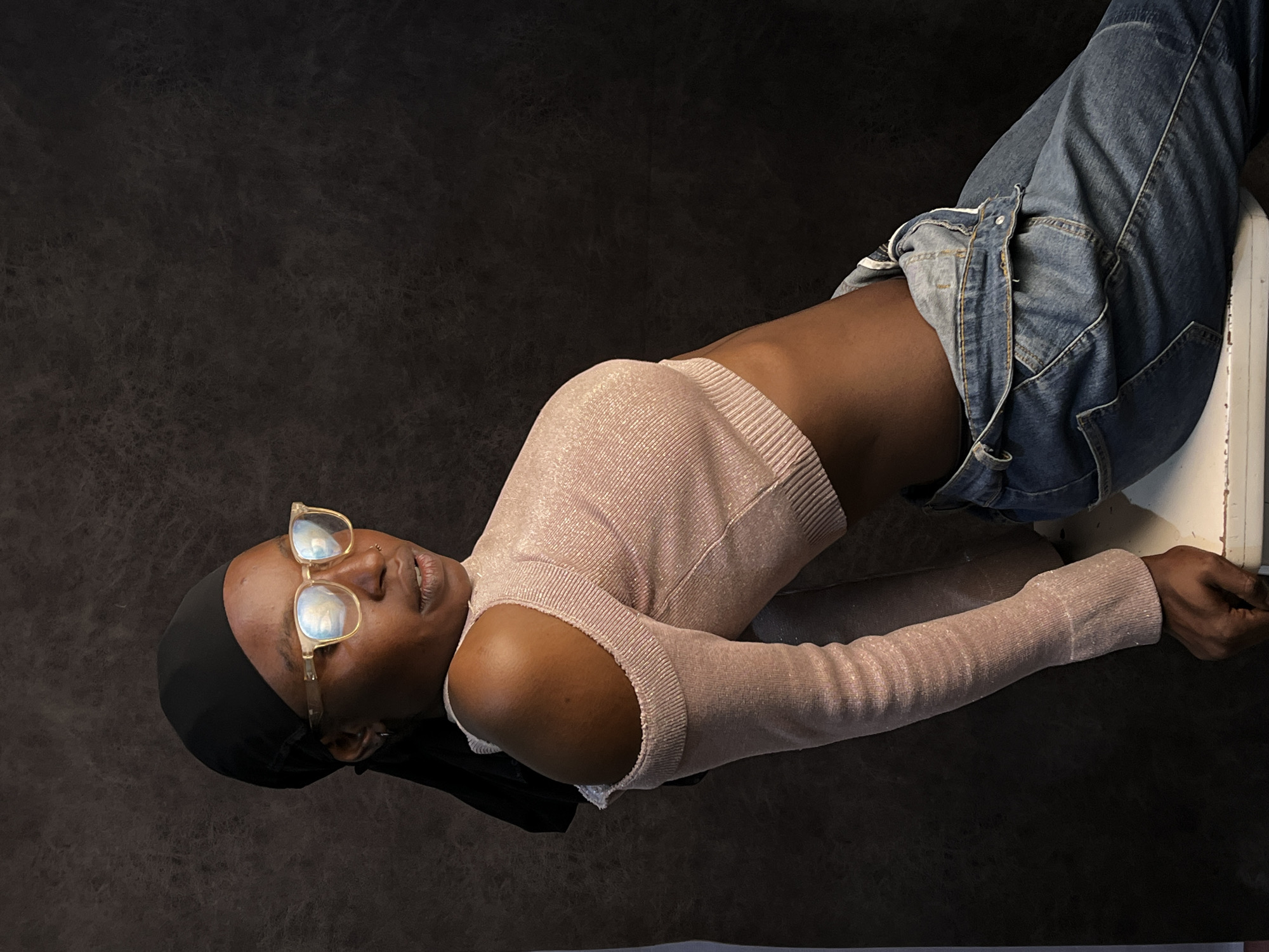 A dark-skinned person with short hair covered by a black cap is lying on their back with their legs elevated on a white object. The person is wearing gold-rimmed glasses, a pink, long-sleeved crop top, and blue jeans. The background is a dark, textured material. The location in the city of Barakin Akawo, Nigeria, is not discernible from the image.