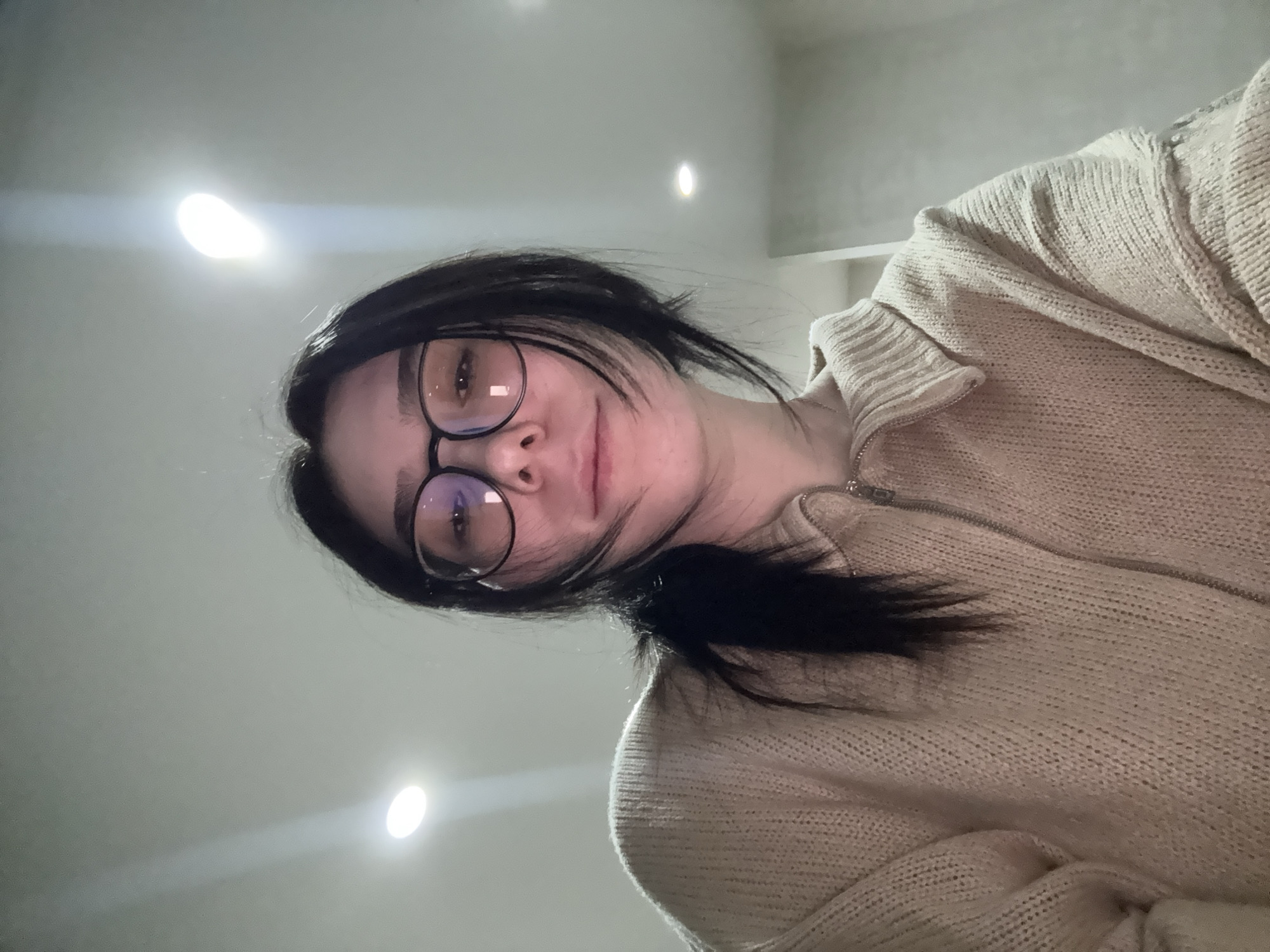 A woman with dark hair pulled into a low ponytail, wearing round glasses and a tan, zip-up sweater, is looking down at the camera. The background is a pale, neutral tone with three diffused, round light sources. The image is taken from a low angle, looking up at the woman. It is not possible to infer the location is Izhevsk, Russia from this image.