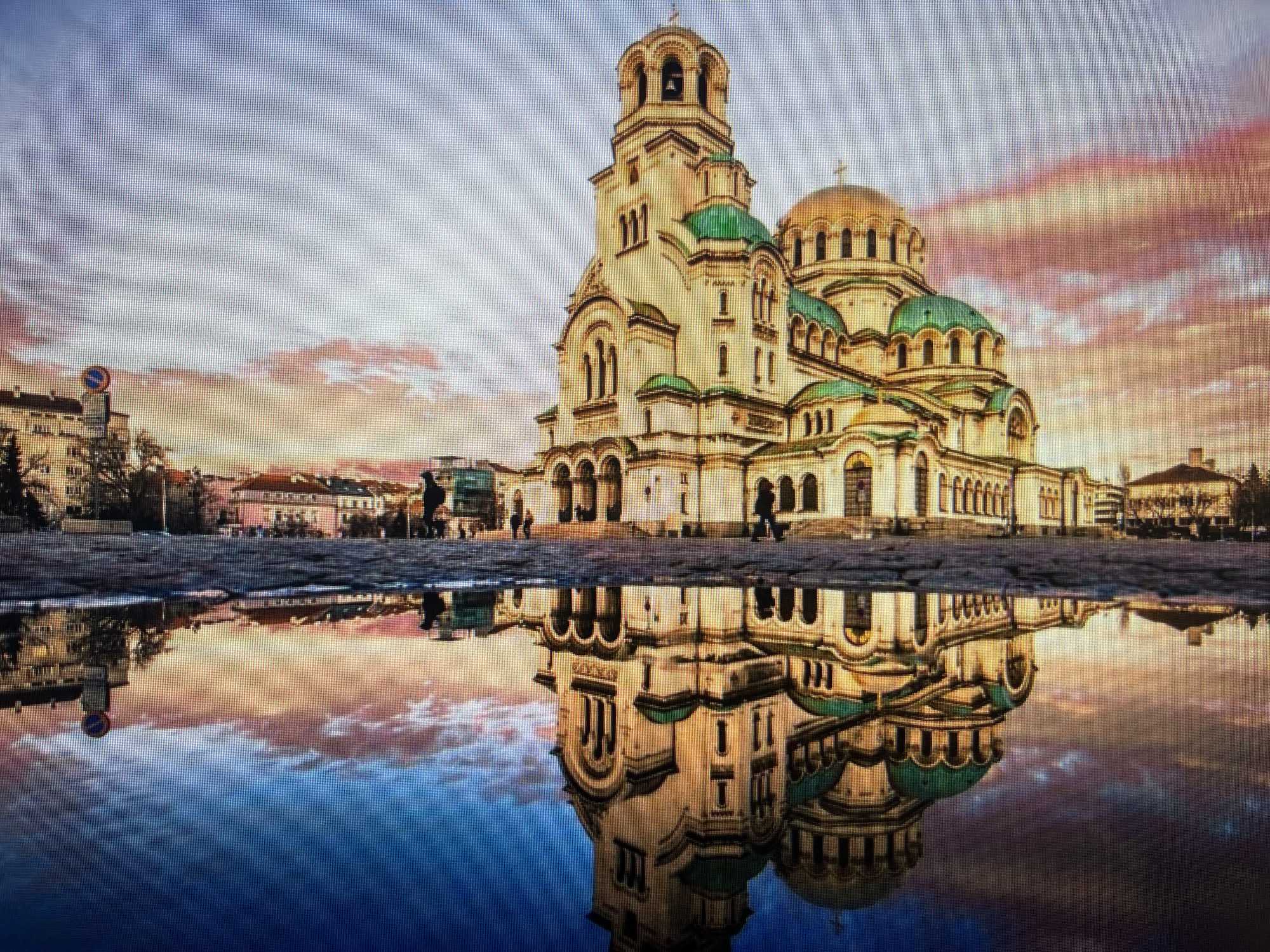 This media file captures a picturesque view of the Alexander Nevsky Cathedral, bathed in the warm, soft light of either sunrise or sunset. The cathedral, with its grand beige facade, prominent bell tower, green domes, and golden cupolas, stands majestically under a sky painted with hues of pink, orange, and blue from the scattered clouds.
A striking feature of the image is the clear and still reflection of the cathedral and the colorful sky in a large puddle or shallow body of water that dominates the foreground. This reflection creates a near-symmetrical composition, doubling the visual impact of the architecture and the vibrant atmosphere.
In the mid-ground, several individuals are seen walking across the cobblestone pavement in front of the cathedral, with their reflections also subtly visible in the water. To the far left, a circular blue road sign with a red diagonal line, typically signifying "No Stopping" or "No Parking," is partially visible near other urban buildings. Additional buildings line the background on both sides of the cathedral.
The scene exudes a sense of tranquility and grandeur, highlighting the architectural beauty against a dramatic natural backdrop. The overall image appears to be a digital photograph, displaying a visible pixel grid, indicative of a screenshot or a photograph of a screen. The setting is an urban environment, with the provided location context being Varna, Bulgaria.
