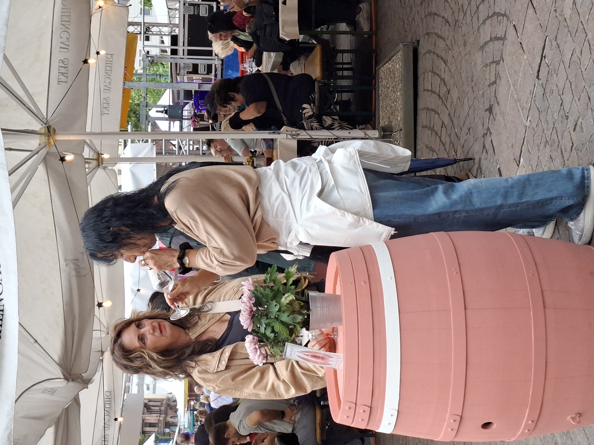 Two women are standing at a table covered by a pink barrel-shaped table, with a potted pink flower in the center. The woman on the left has long, wavy brown hair and is wearing a tan jacket over a dark top. She is looking to the left and holding a glass of red wine. The woman on the right has long, dark hair and is wearing a tan sweater over a white shirt and blue jeans. She is holding a glass of white wine to her lips and looking to her right. Behind them, a white tent is set up with string lights. People are seated at tables in the background, with a stage and sound equipment visible further back. The ground is paved with cobblestones. The scene appears to be an outdoor event, likely a wine festival, possibly in the city of Wiesbaden, Germany, given the presence of a tent with what appears to be German text visible on it and a general atmosphere typical of European outdoor markets or festivals.
