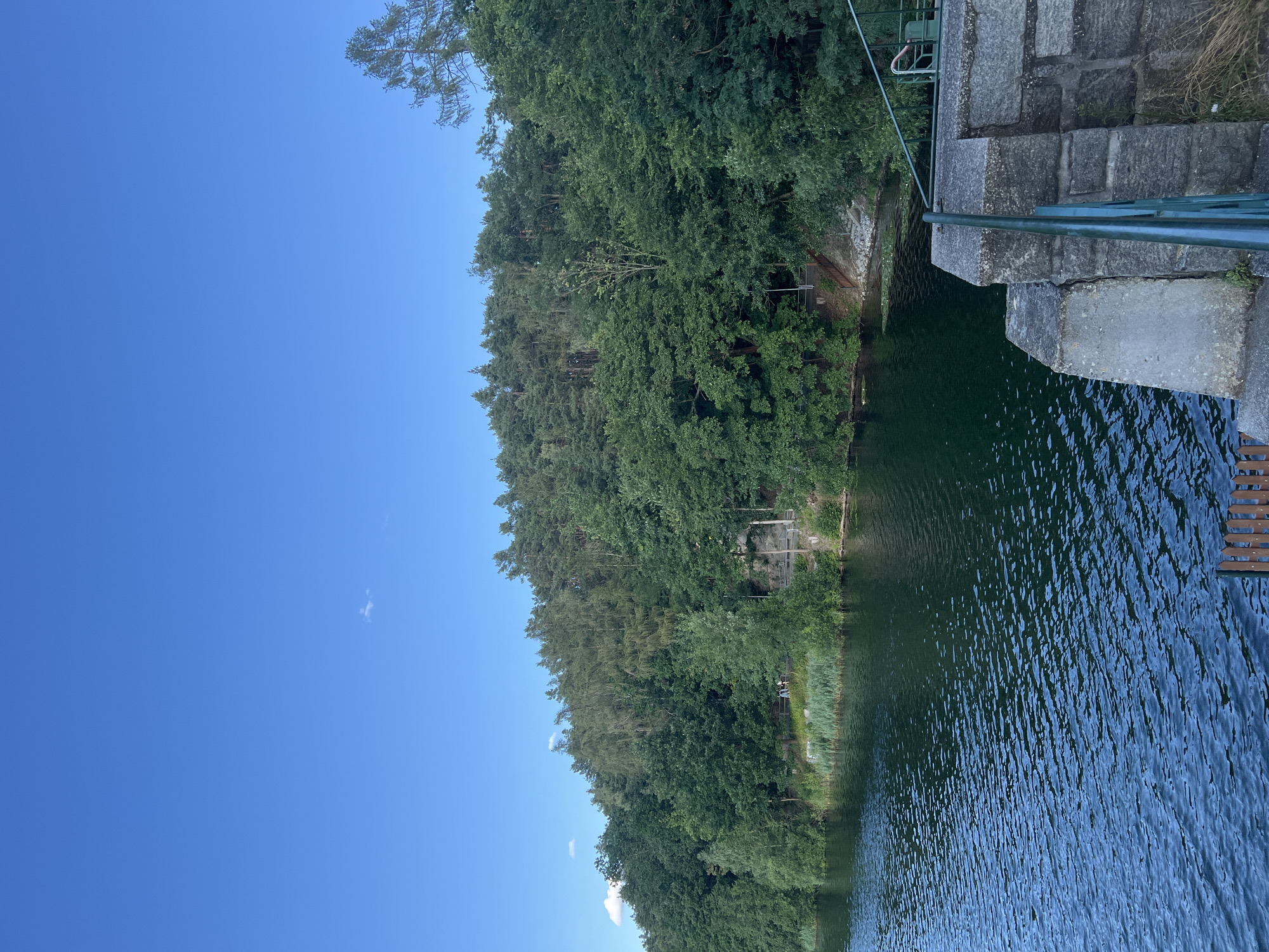 A wide shot captures a tranquil lake bordered by a dense, green forest under a clear blue sky. The water, dark and rippling, reflects the surrounding trees. In the foreground, a stone structure with a green railing juts into the scene, hinting at a dam or embankment. A wooden platform is partially visible at the bottom left. Along the opposite shore, a narrow pathway winds through the trees, where a few people can be seen walking. The image depicts a serene natural landscape with man-made elements integrated into the environment.