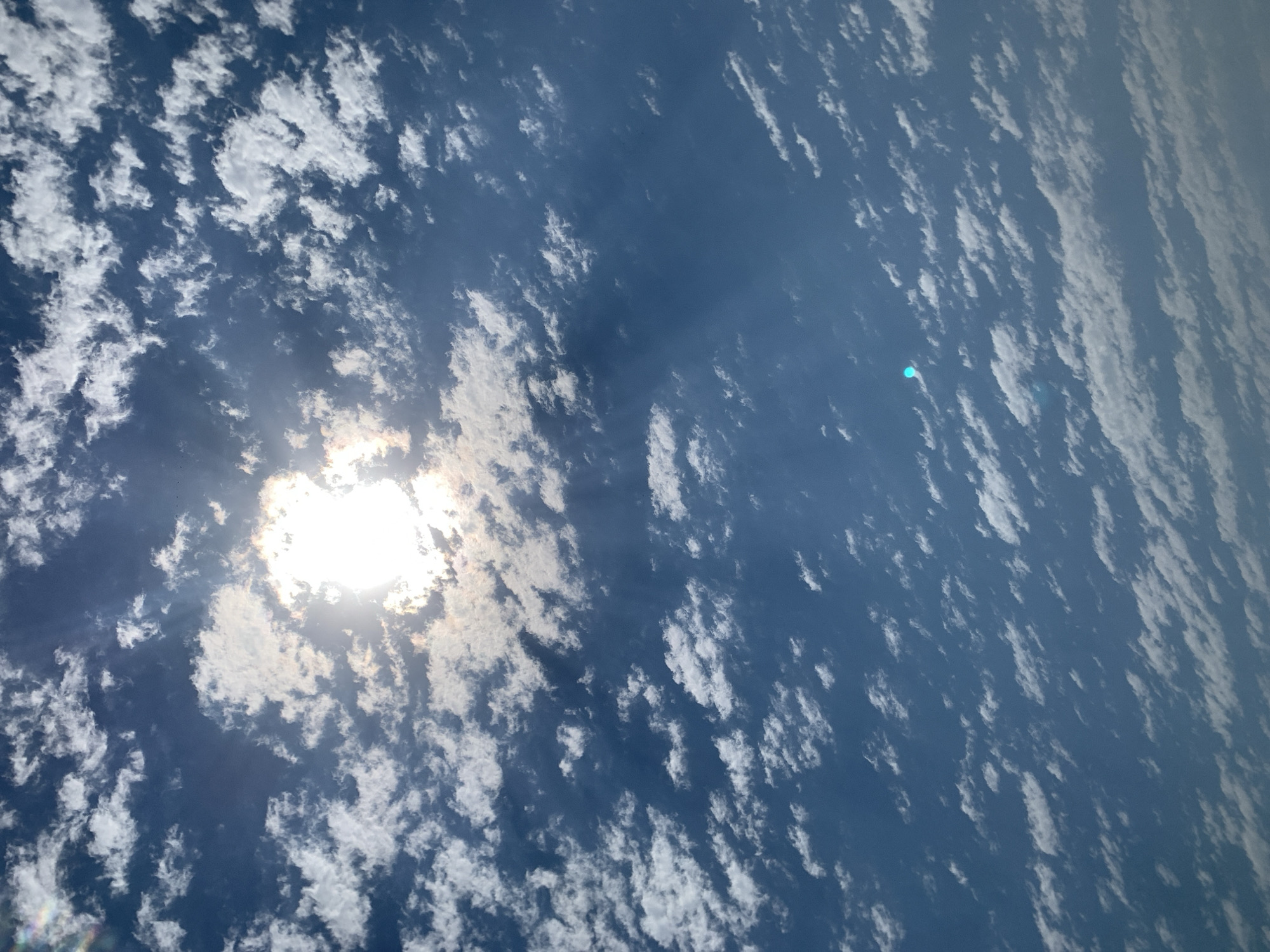 The image shows a bright sun partially obscured by scattered white clouds against a deep blue sky. Sunlight rays are visible. There is a lens flare in the bottom center of the image, appearing as a small blue and green spot. The clouds are distributed across the frame, with some denser patches and others more wispy. The scene is devoid of any discernible landmarks, people, or man-made objects that would indicate a specific location within Khazar Raion, Azerbaijan, or any other urban setting. The focus is solely on the sky and atmospheric conditions.