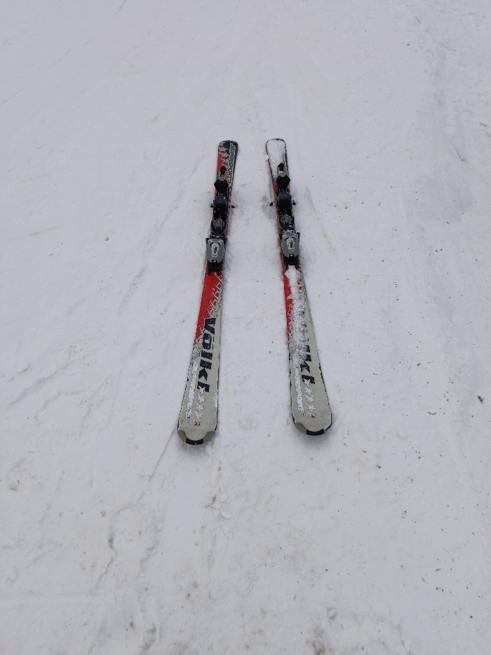 The image captures a pair of skis lying on a snow-covered ground, likely a ski slope or a snowy mountain environment. The skis are positioned parallel to each other, angled slightly from the upper right to the lower left of the frame.
Both skis are predominantly red and white/silver in color with black tips and tails. They feature black bindings mounted towards the center. The tops of the skis are lightly dusted with loose snow, suggesting recent use or current exposure to snow. The snow on the ground appears somewhat packed and has faint textures and minor depressions, indicating it's not freshly fallen and may have been skied on or walked over.
Text visible on the left ski, oriented vertically along its length, reads "VOLKL" in white lettering on a red background. Further up on the same ski, closer to the bindings, the word "SUPERSport" is also visible. The skis also display decorative star-like patterns and other graphic elements in white against the red and silver areas. The bindings are black with silver-colored metallic components.
The scene conveys a sense of a winter sports activity, with the skis at rest on a cold, snowy day. The lighting appears even, suggesting overcast conditions or a uniform light source.