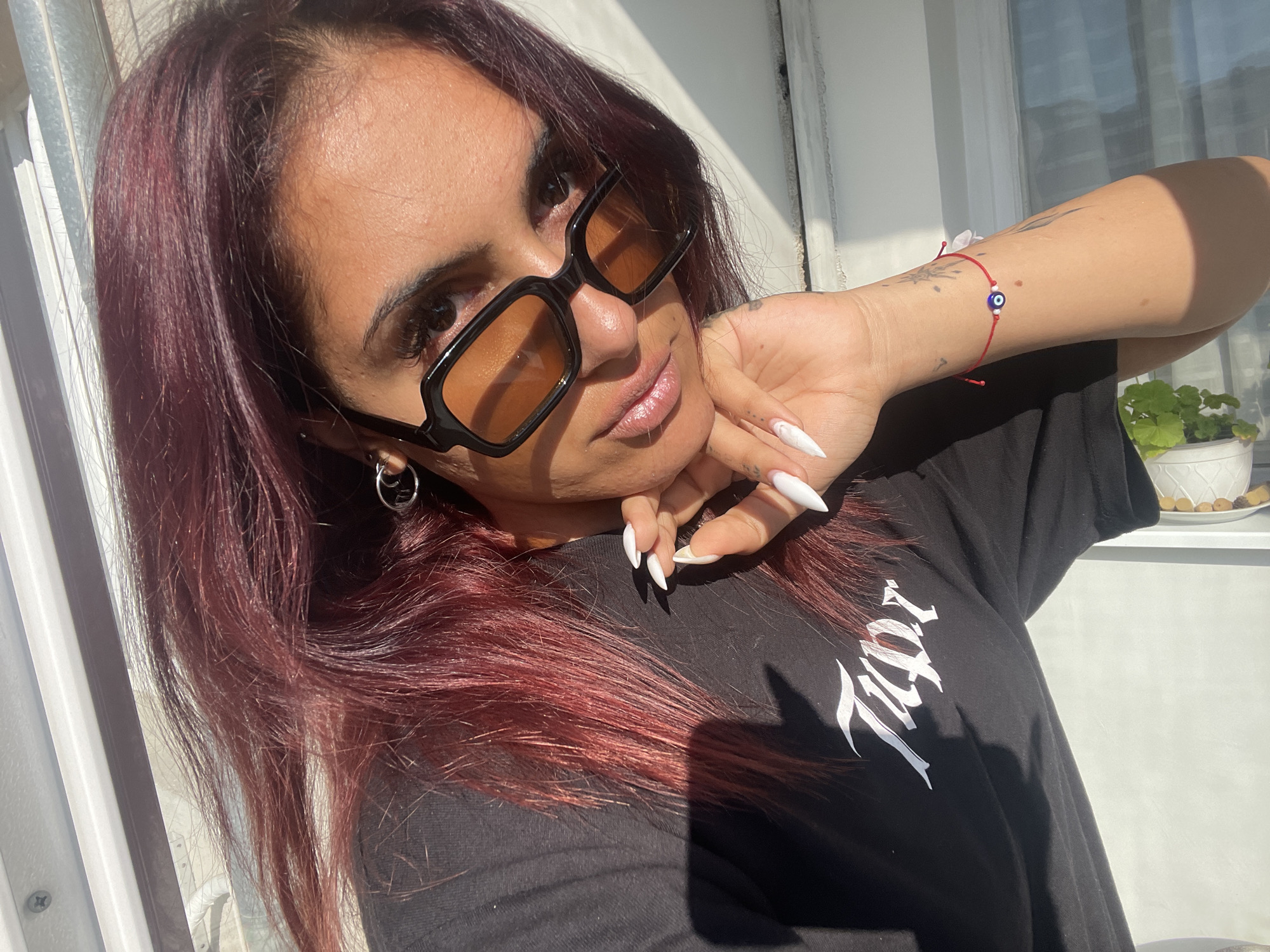 A woman with auburn hair, wearing black-framed sunglasses with orange lenses and a black t-shirt with white lettering, poses in an outdoor location. She is wearing a silver hoop earring in her left ear and a red string bracelet with a blue eye charm on her right wrist. Her nails are long, white, and pointed. In the background, there is a window with a plant in a white pot on the windowsill. The location in the city of Unknown, Unknown is not determinable.