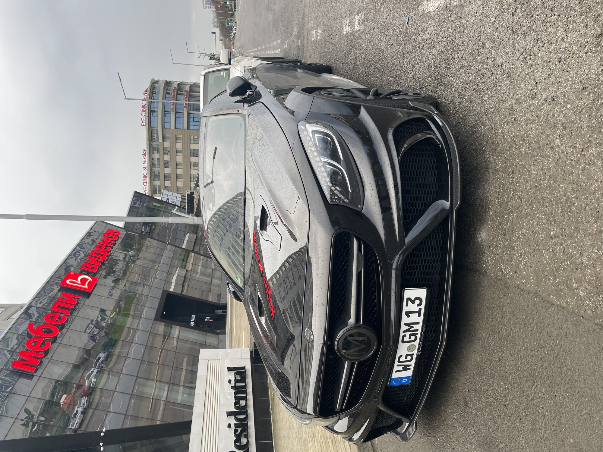 The image is a close-up shot of the front of a black sports car parked on a wet city street. The car features a custom grill and license plate that reads "WG GM 13," indicating it is registered in Germany. The background includes a modern building with a prominent red sign reading "Мебели Виденов" (Furniture Videnov) in Cyrillic. Next to it, the sign of "Residential" in English is visible. Across the street, a taller building with a sign "EYE CUNIC St. Nikolay" is also present. The scene suggests an urban environment in Burgas, Bulgaria.