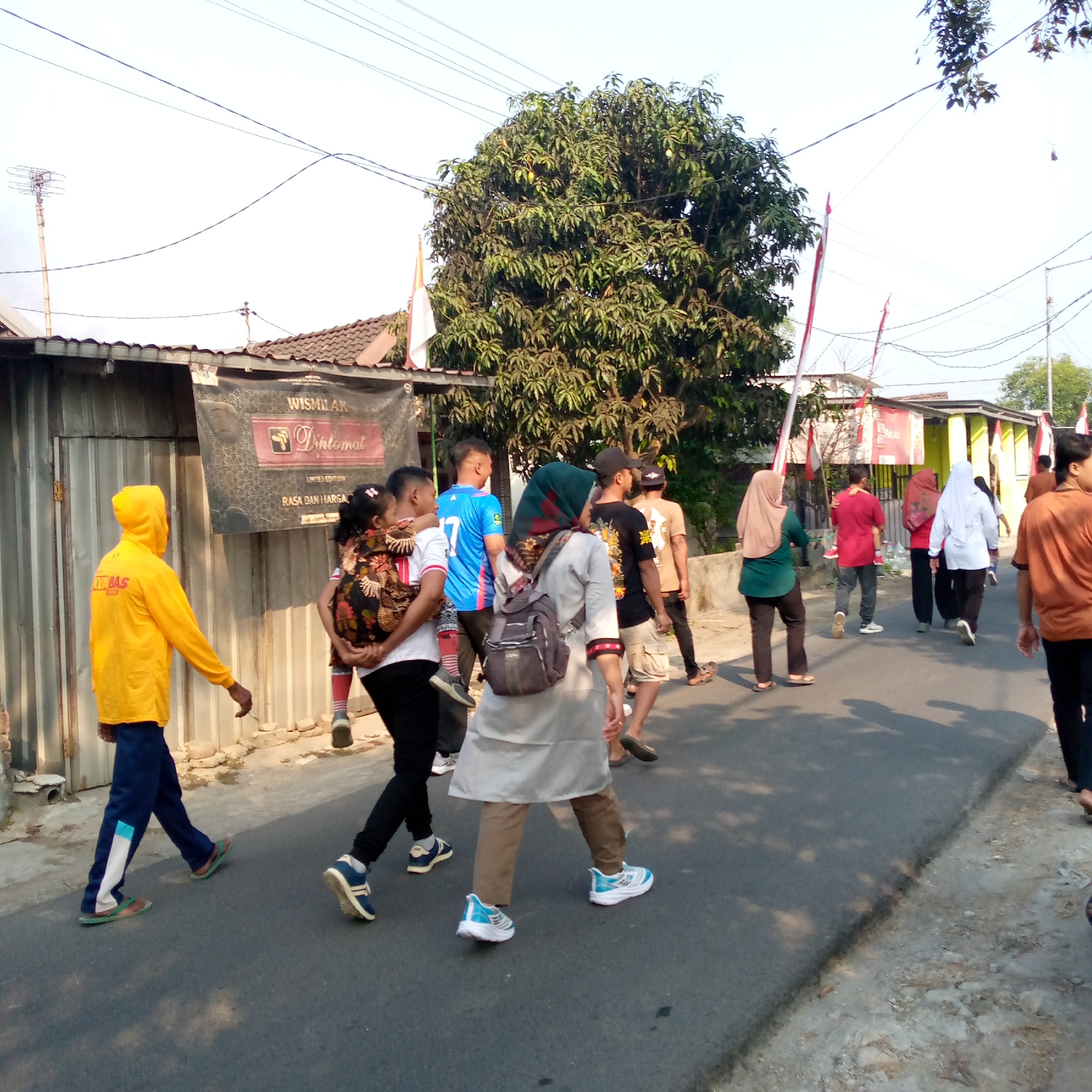 In Tambakagung, Indonesia, a group of people are walking on a paved road with a tree and buildings in the background. The scene is captured during daylight with a bright sky. In the foreground, a person wearing a yellow hooded jacket and blue pants walks away from the camera. To their left, a woman in a gray outfit with a headscarf and backpack walks forward. Further back, several other individuals are walking in the same direction, some in casual attire and others in headscarves. Indonesian flags, red and white, are visible on poles along the roadside. The buildings appear to be simple structures with metal siding and tiled roofs. The overall impression is of a casual gathering or procession on a street in a residential or commercial area.