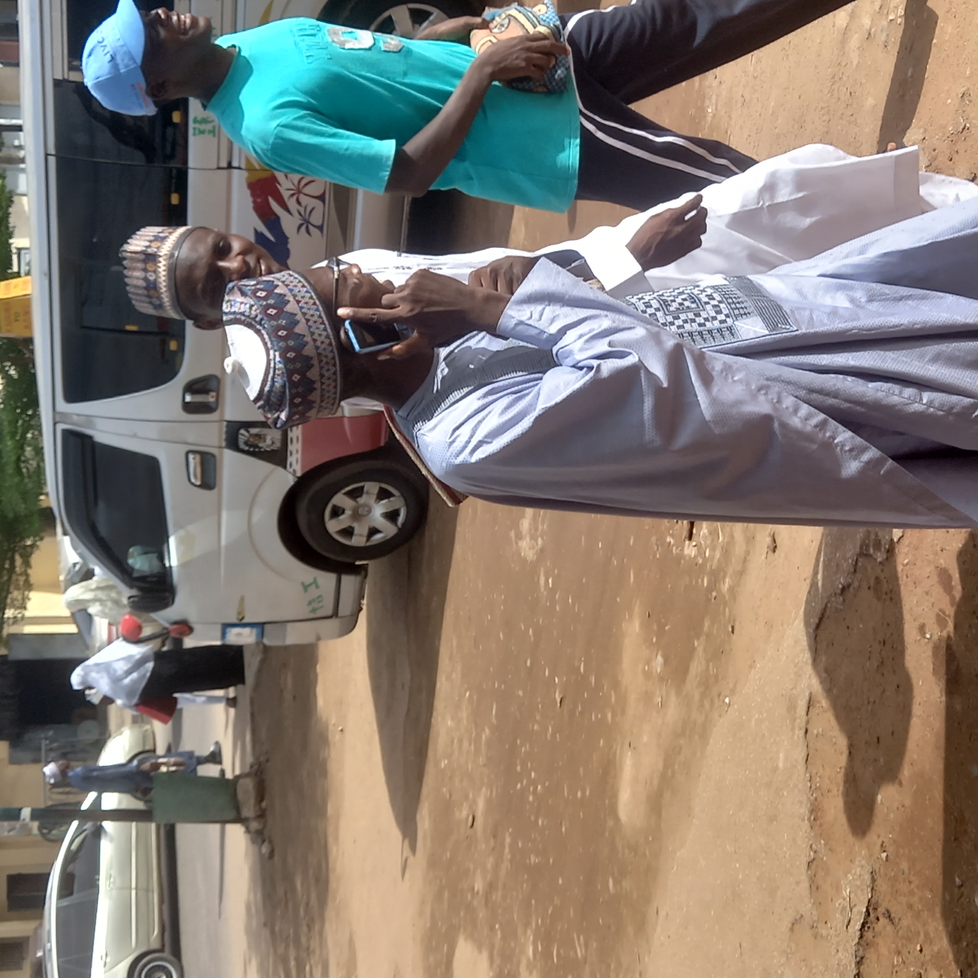 In Dadi Riba, Nigeria, the image shows two men standing outdoors on a partially paved street. One man, wearing a blue traditional cap and a light blue robe, is speaking on a smartphone. The other man, wearing a blue baseball cap and a turquoise shirt, stands nearby. Behind them is a white van with red and black accents. Other people and parked vehicles are visible in the background. The ground is a mix of pavement and dirt.