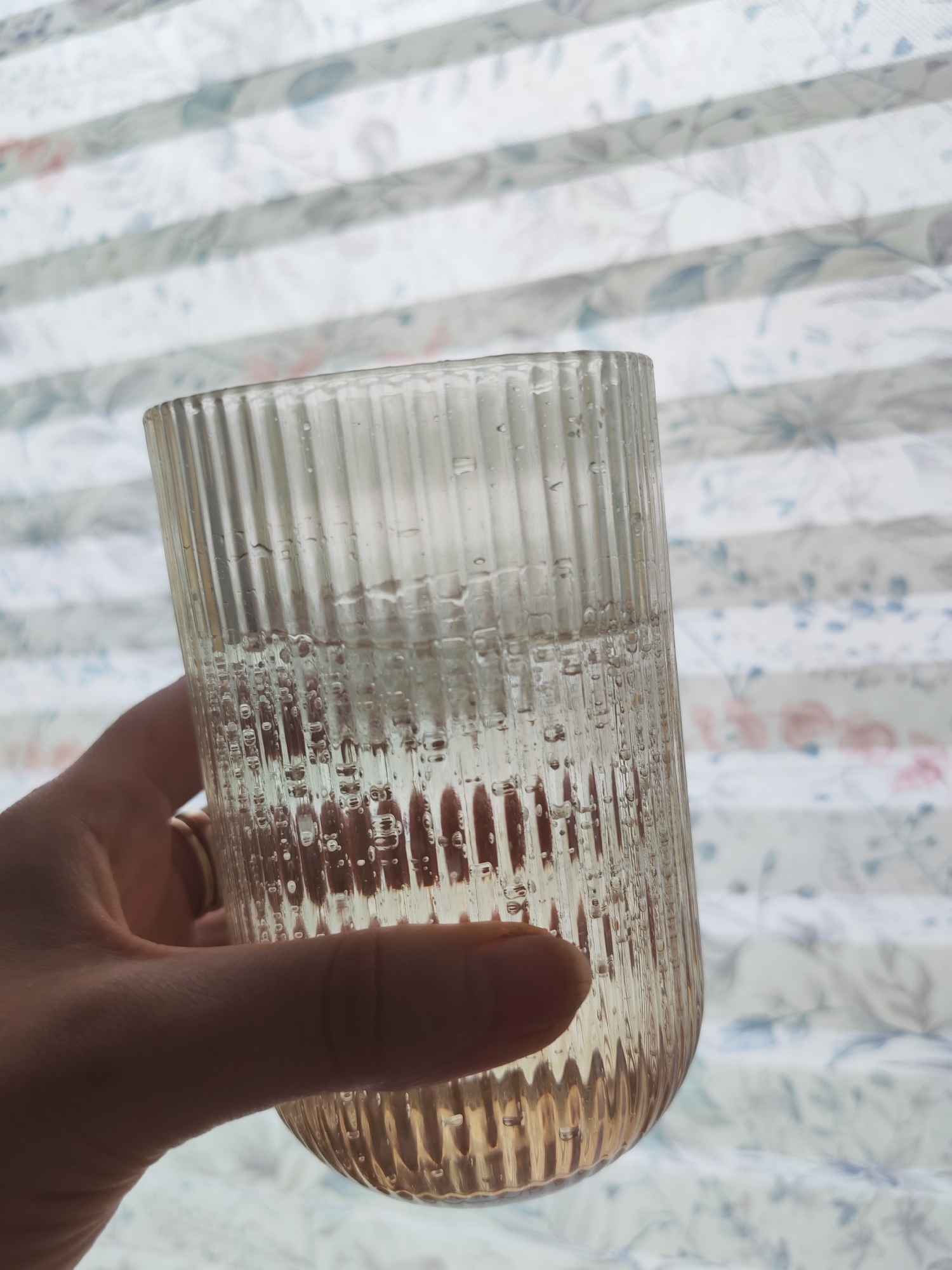 A close-up shot shows a hand holding a ribbed glass filled with a clear liquid, likely water, with small bubbles visible inside. The glass is positioned in front of a window with white blinds that have a subtle floral pattern. The lighting suggests it is daytime. The origin of the image is confirmed to be Izhevsk, Russia.