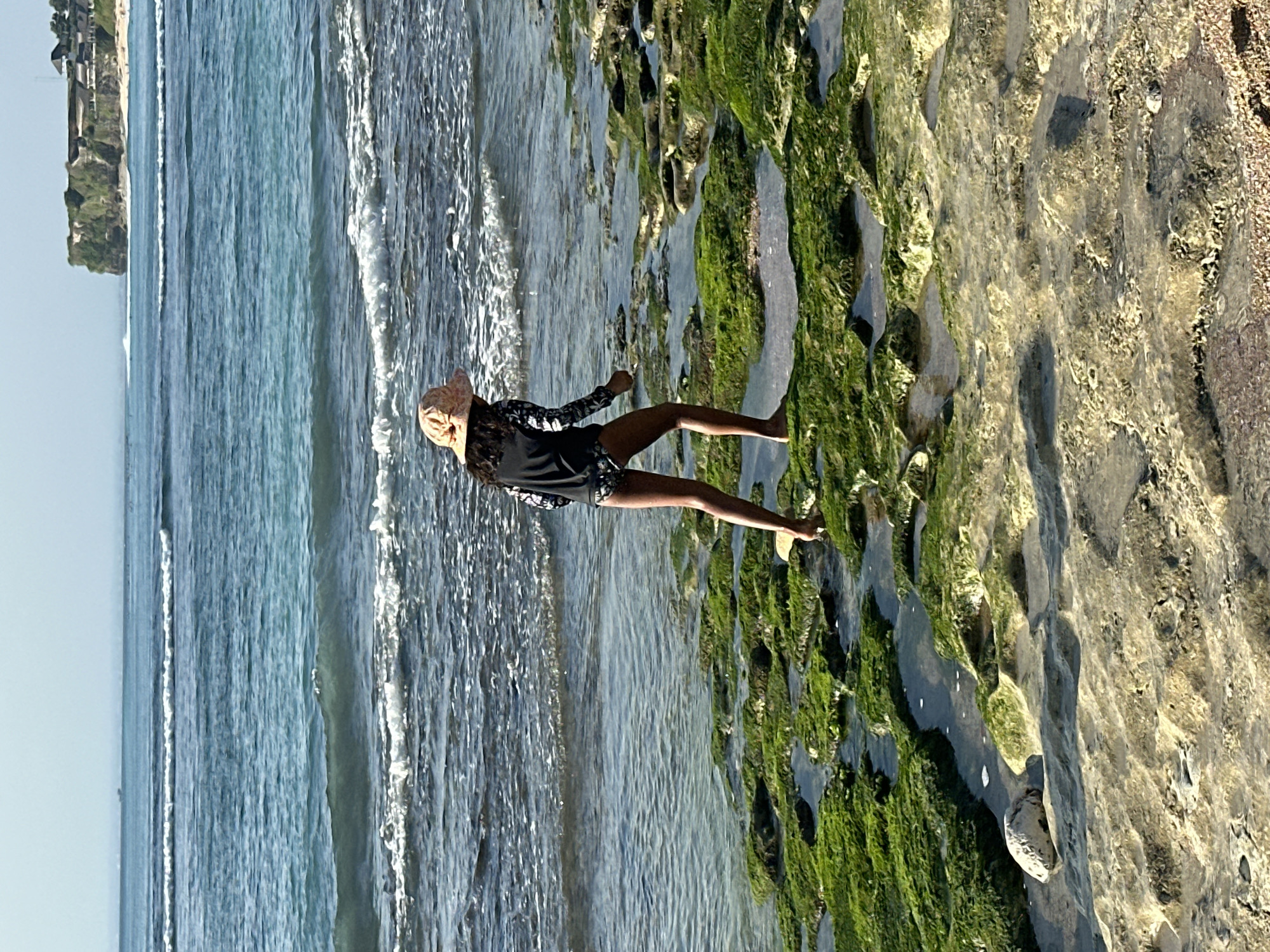 The image shows a young person wearing a swimsuit and a hat walking on a rocky, algae-covered shoreline in Kuta Selatan, Indonesia. The ocean is visible in the background with gentle waves. Cliffs and buildings can be seen in the distance along the coastline. The lighting suggests it is daytime.