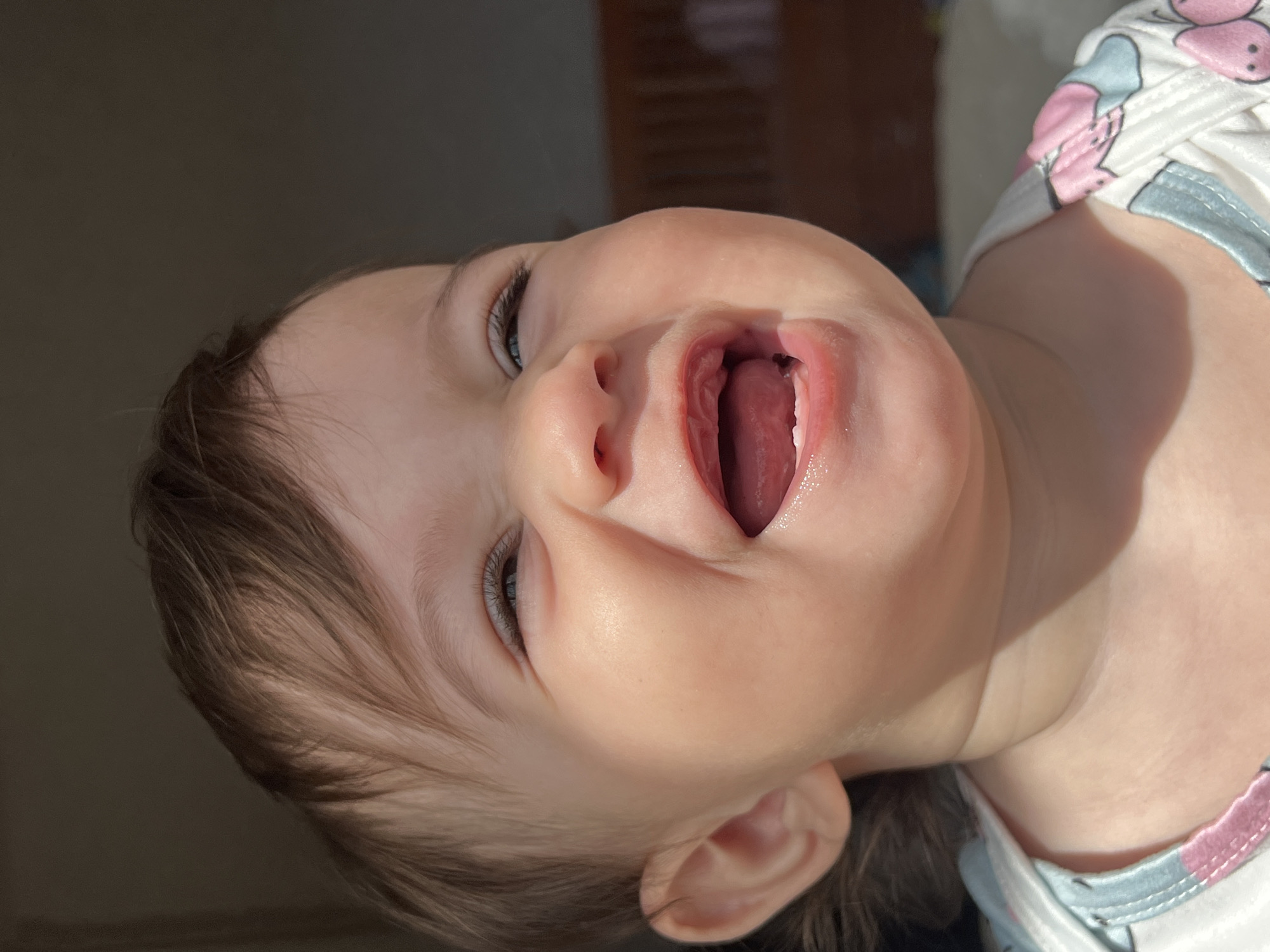 A baby lies on their back, head tilted back and mouth open wide, revealing their tongue. The baby has fair skin, brown hair, and blue eyes. They are wearing a light-colored shirt with pink and gray floral patterns. The background is blurred, but appears to be indoors with some light coming in from the left. The location in the city of Elena, Bulgaria is not visually discernible.