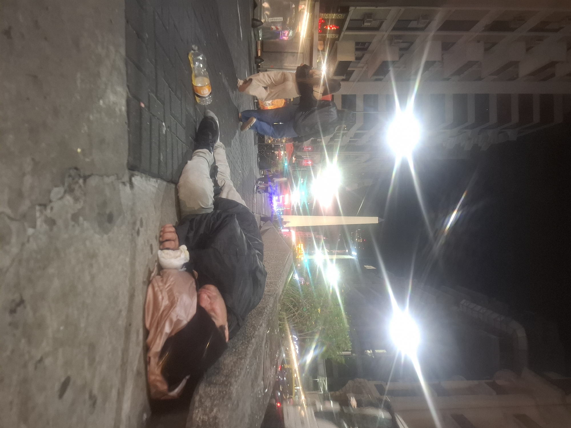 The image appears to be taken at night on a street in Buenos Aires, Argentina. A person is lying on the sidewalk next to a curb. The person is wearing light colored pants, black shoes, a black jacket and a light colored shirt. They are lying on their side with a pink bag. An empty plastic bottle sits on the pavement near the person's feet. In the background, other people are walking along the sidewalk. The street is lined with buildings and bright streetlights.