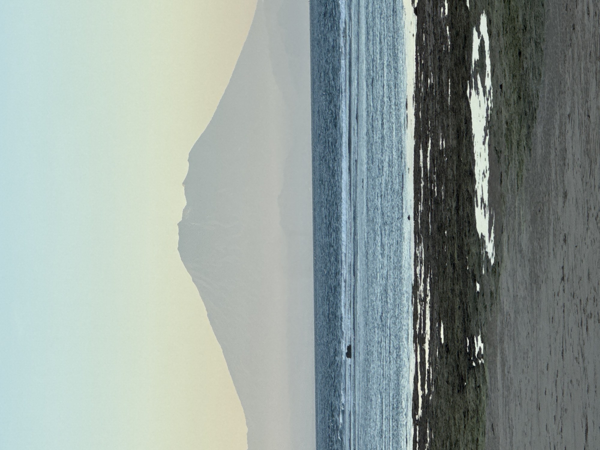 The image depicts a landscape with a mountain in the background, partially obscured by haze. A body of water, possibly the ocean, is visible, with gentle waves. The foreground appears to be a shoreline with dark vegetation and wet sand. The location is likely in Kuta Selatan, Indonesia, given the tropical landscape and coastal environment. No people are visible.