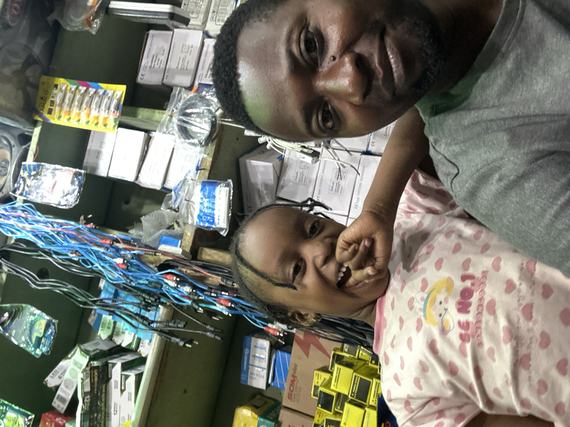 An indoor selfie features an adult male and a young girl positioned centrally, set in Narayi, Nigeria. The male, on the right, has short dark hair and a beard, wearing a grey t-shirt, and looks directly at the camera with a slight smile. The girl, on the left, has braided hair and is captured mid-laugh, with her mouth open and one hand near her chin. She wears a pink t-shirt with numerous small pink heart patterns and a cartoon character design featuring "SE NO.1". The background reveals a cluttered environment, likely a shop or storage area, with wooden shelves stacked with various products. Observable items include numerous cardboard boxes, some labeled "LED", "SOLAROSYS", and "SOM elec", suggesting electronic or lighting supplies. Bundles of electrical cables, predominantly blue and black with red and white connectors, hang prominently. A yellow card displaying "AMACCI" batteries is also visible on a shelf. Other assorted packaged goods and components are scattered throughout the shelves.