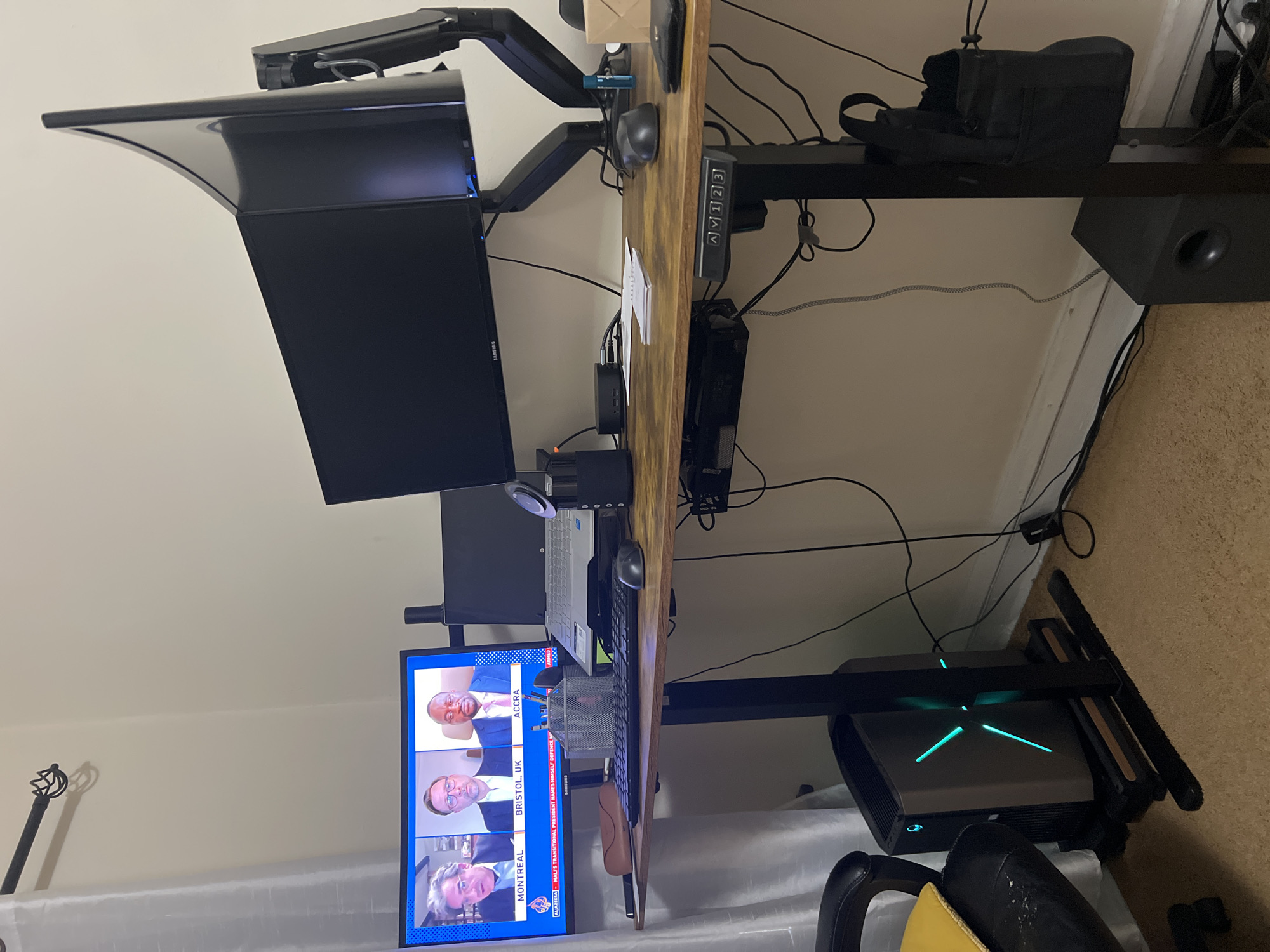 Here's a detailed description of the image:
**Scene Overview:** The image shows a workspace setup, likely inside a home office. The dominant features are the computer equipment and a wooden-topped desk.
**Objects:**
* **Computer:**
* A computer monitor is displaying a news broadcast on Al Jazeera. The news ticker references "Mali's Transitional President".
* Multiple monitors are stacked above the visible display.
* A laptop is open on the desk.
* A keyboard and mouse are present.
* A desktop computer tower is visible on the floor, featuring a green "X" design.
* **Desk:** The desk has a wooden top and a black frame. A control panel is on the right side of the desk.
* **Audio:** Speakers are on top of the desk.
* **Accessories:** An eyeglasses case and writing instruments are on the desk.
* A monitor arm supports the displays.
**Environment:**
* The wall behind the desk is painted white.
* The floor appears to be carpeted.
* Cables and wires are visible, running along the desk and down to the floor.
* The presence of a curtain indicates a window.
* The image does not explicitly state the city of Chicago, United States.
* The image is taken indoors.
**People:** There are four people in boxes displaying on the lower screen. The locations of the people are Montreal, Bristol (UK), Accra, and another not visible in the cropped image.