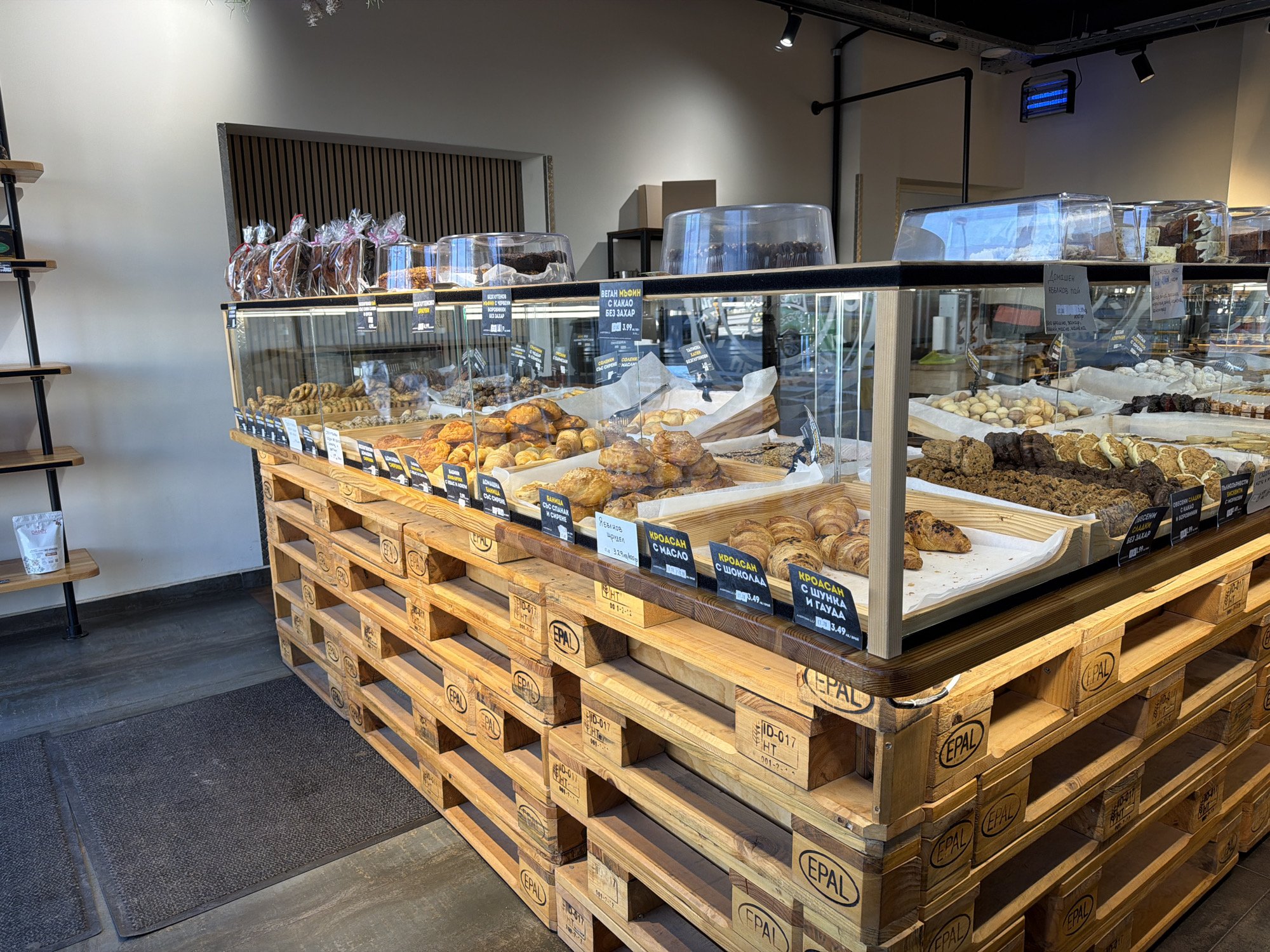 The image shows a bakery display in Sofia, Bulgaria. A large display case filled with pastries sits atop a structure built of wooden pallets, several layers high. The pallets have "EPAL" and "ID-017 HT" markings. In the display case are various baked goods like croissants, muffins, and other pastries, each with small signs indicating the item name and price in Bulgarian currency. The labels include "BAHMCA C CIPEHE," "BAHMCA C CПАHAК," "Ябълков Щрудел," "КРОАСАН C МАСЛО," "КРОАСАН С ШОКОЛАД," "КРОАСАН C ШУНКА И ГАУДА," and "ВЕГАН МЪФИН С KAKAO БЕЗ ЗАХАР." Some items are under clear dome-shaped covers.
Behind the display case, there's additional merchandise. The bakery appears to be indoors. To the left, there is a dark doormat on the floor and a tall shelving unit. The background includes walls of various colors, with a section featuring vertical wooden slats. The overall lighting is bright. A blue-colored light fixture is visible in the upper-right corner of the image.