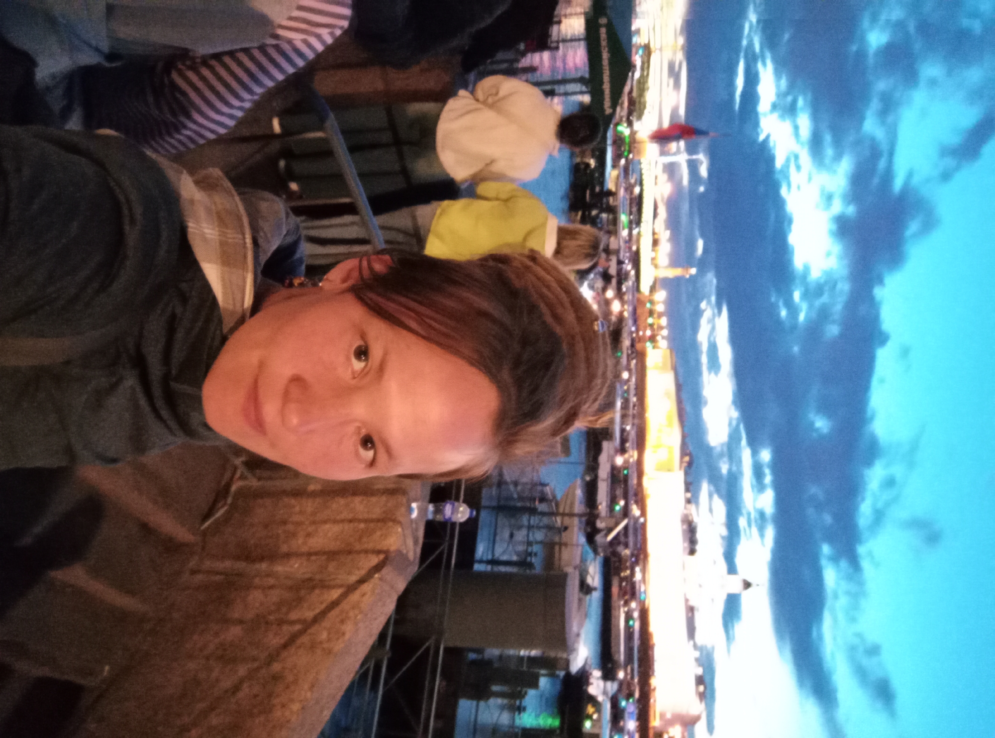 An eye-level, medium shot captures a woman taking a selfie on a quay at dusk. The woman is in the foreground, slightly off-center to the right. She has light skin, auburn hair styled into dreadlocks, and is wearing a dark blue hooded jacket. Her right eye is visible, and she has a faint smile.
Behind her, two women stand near a railing, facing away from the camera. The woman on the left wears a light-colored jacket, and the woman on the right wears a bright yellow hooded jacket. Both are looking out at the water and the cityscape.
The background reveals a waterfront scene with boats docked and illuminated buildings. The sky is a dramatic twilight blue with streaks of dark clouds and some bright patches. A Russian flag flies on a pole to the left of the frame. The overall atmosphere is cool and dusky.