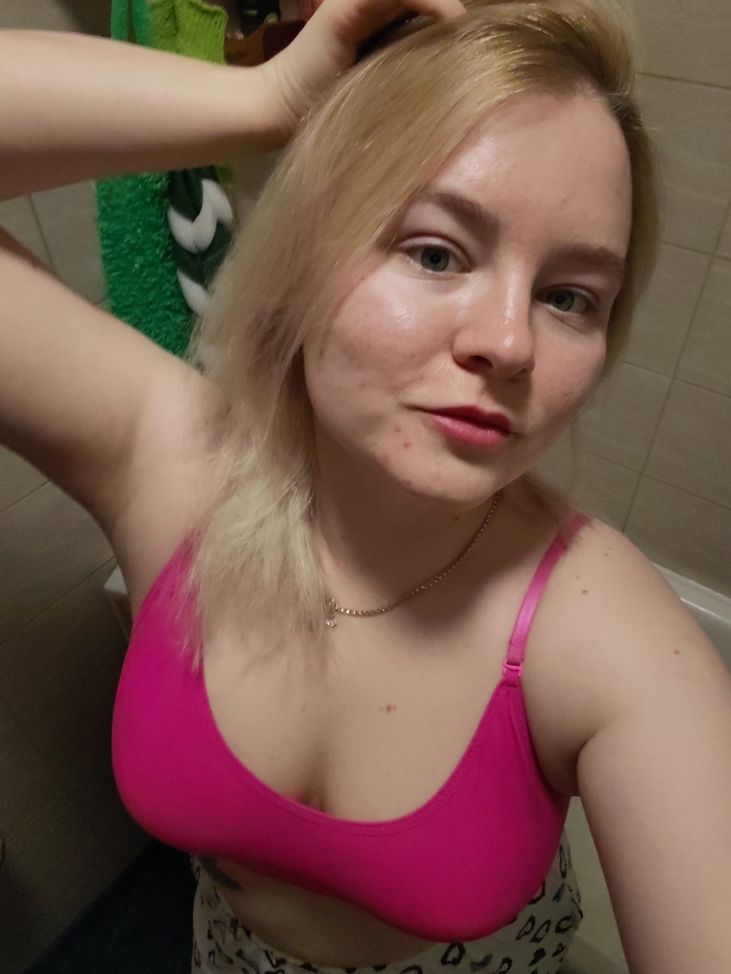 A woman is in a bathroom. She is wearing a pink bra and patterned pants. She has blonde hair and fair skin. Her right arm is raised and her hand is touching her hair. A green and white towel hangs on the wall behind her. The background shows beige tiles. The image was taken in a bathroom but the exact location in Izhevsk, Russia, cannot be determined from the information available.