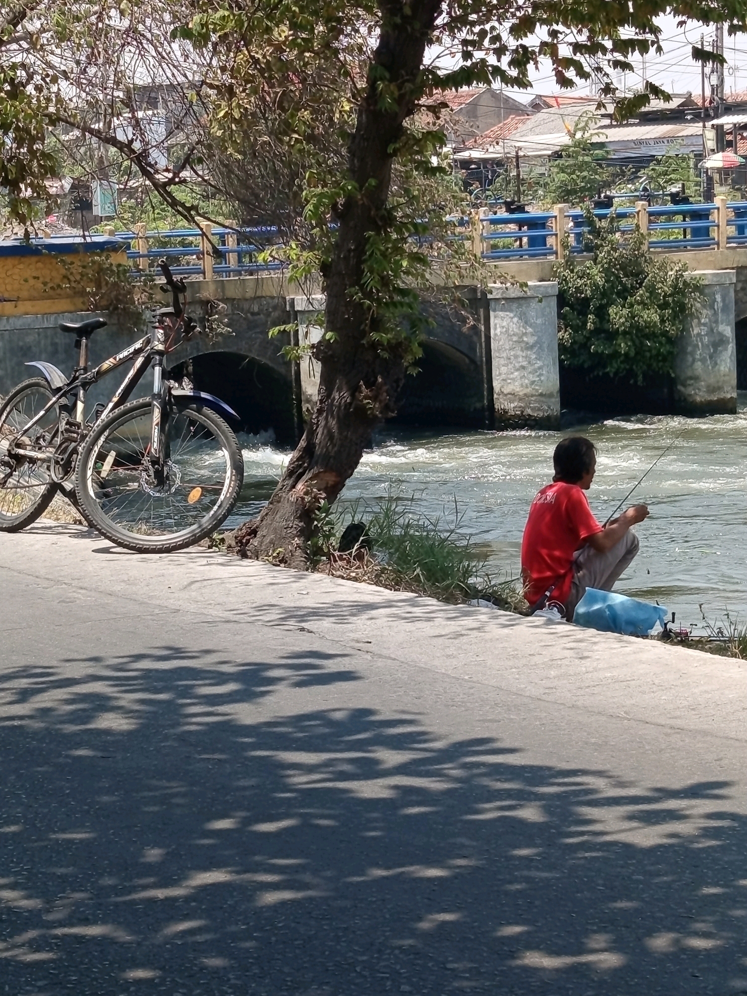 A person is fishing by the side of a river, with two bicycles parked nearby. The scene is likely in the city of Karawang, Indonesia, given the presence of a bridge with blue railings and the general architectural style of the buildings in the background, which appear to be a mix of residential and commercial structures. The river is flowing, and there are some trees and vegetation along its banks. The person is crouched down, holding a fishing rod, and appears to be focused on their activity. The bicycles are parked on a concrete path next to the river, and a blue bag is placed on the ground beside the person. The overall impression is of a quiet, everyday scene in an urban environment.