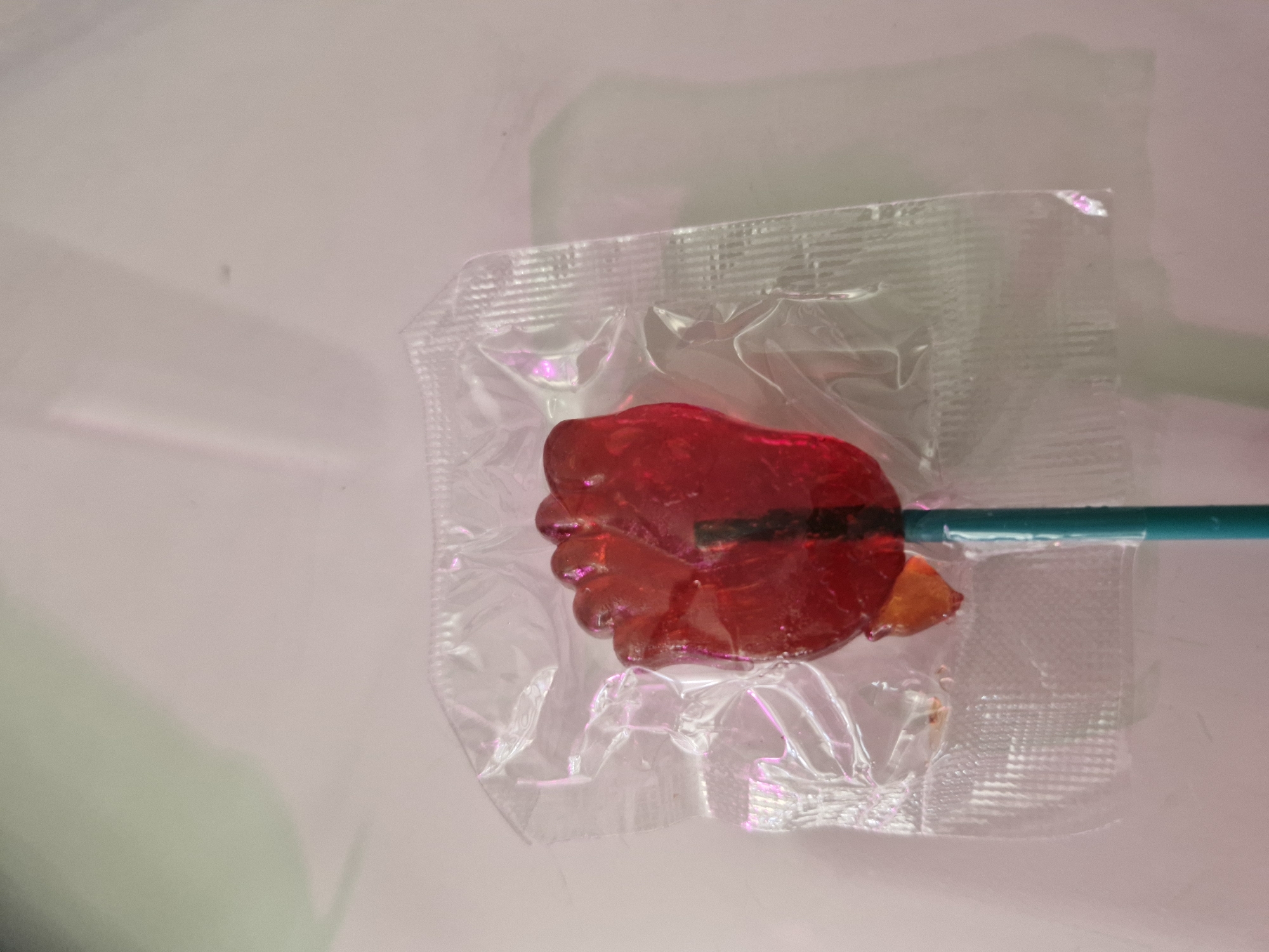 A red, foot-shaped lollipop is centered in the frame, partially enclosed in clear plastic packaging. The lollipop has five distinct toe shapes and a dark stick. An orange, irregular piece of candy is visible at the bottom left of the lollipop, also within the packaging. The lollipop stick is teal and extends downwards out of the frame. The background is a pale pink surface with soft shadows casting from the object, suggesting an indoor setting. There is no indication of the city of Kazan, Russia in the image.