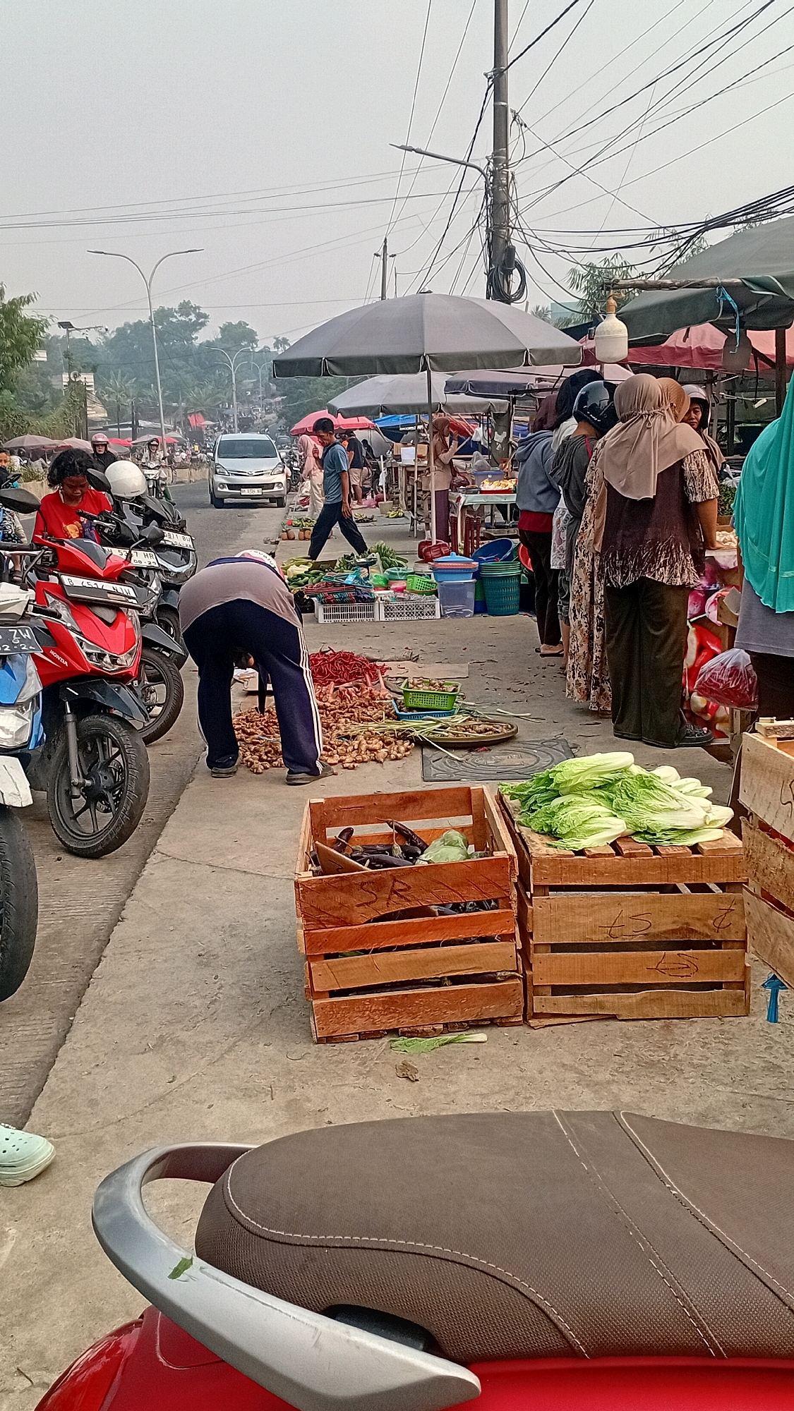 The image depicts a bustling street market scene in Jambi, Indonesia. In the foreground, a red motorcycle seat is visible, suggesting a mode of transportation. To the left, several other motorcycles are parked along a sidewalk. A person wearing a grey shirt and dark pants is bending down to examine goods on the ground, which appear to be ginger or similar root vegetables. The sidewalk is lined with various stalls, some covered by umbrellas, selling produce. In the background, a white car is driving on a street, and more people are seen walking and browsing at the market. The environment suggests an outdoor market area with a mix of pedestrian and vehicular traffic, characteristic of a busy commercial street in Jambi.