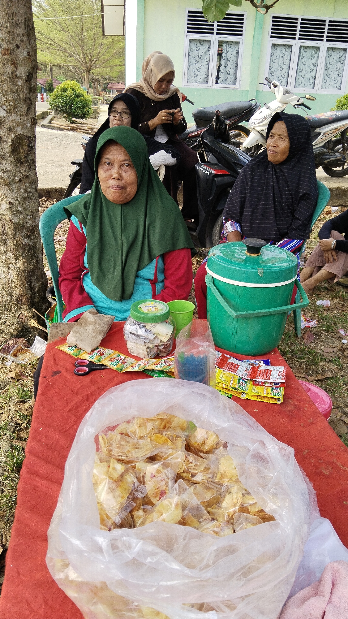 In Margatunggal, Indonesia, four women are gathered near a light green building. The woman in the foreground, wearing a green hijab and a red and turquoise outfit, is seated at a red-clothed table and looking forward. To her left, a woman in a black hijab and glasses is also seated. Behind them, two other women are present, one in a light brown hijab and the other in a black hijab. They are positioned near motorcycles. The table displays various items, including a large green bucket, a jar filled with cash and papers, packets of snacks, scissors, and a bag of chips. The surrounding environment appears to be outdoors, with trees and dry foliage visible.