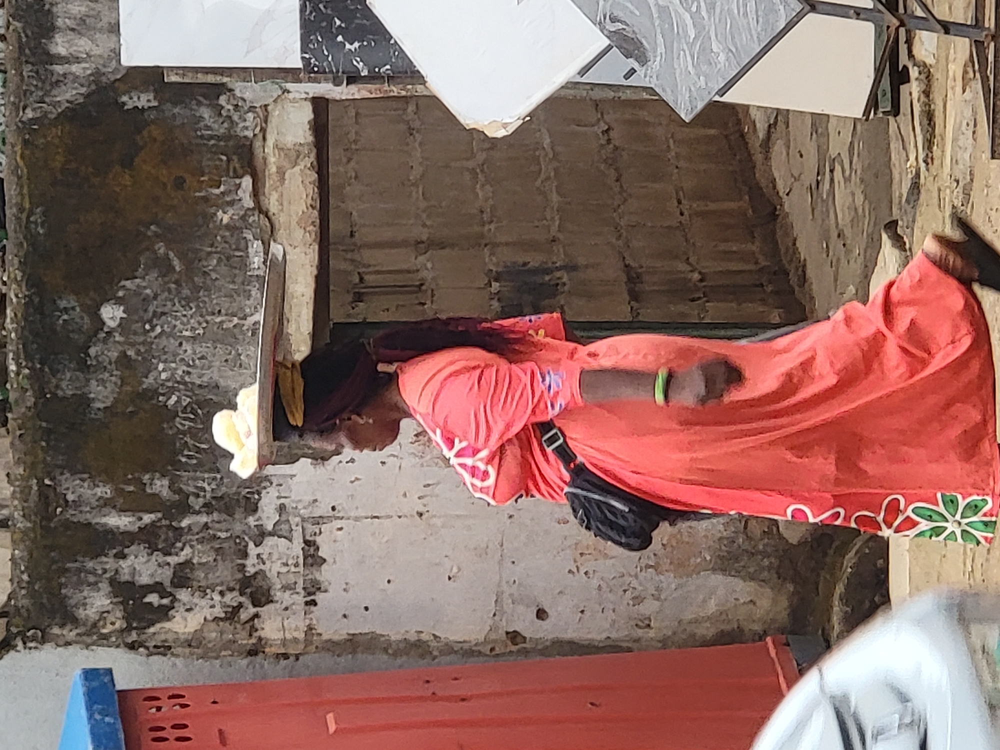 The image, when viewed in its correct upright orientation, captures a person, likely a woman, standing outdoors in what appears to be a commercial or market setting in Maiburiji, Nigeria. She is dressed in a vibrant coral-colored or bright orange long gown, featuring decorative green, red, and white floral-like patterns along its hem. A dark fanny pack is worn across her body, and a bright green bracelet adorns her right wrist. Her head is partially covered by a yellow or gold headscarf, with dark hair visible beneath. She is looking downwards, her expression not clearly discernible.
The background consists of weathered and discolored walls, possibly concrete or plaster, displaying extensive signs of wear, peeling paint, and uneven textures. Above the person, several square tiles with white, black, and grey marble-like designs are displayed, mounted on a structure, suggesting a shop or stall dealing in building materials or home finishes. Behind these displayed tiles, a darker, recessed area hints at an interior space. In the lower foreground, a partial view of a red object with a blue top, possibly a vending machine or a utility stand, is visible. The lighting is soft and diffused, indicating it is daytime without direct sunlight. There is no visible text in the image.