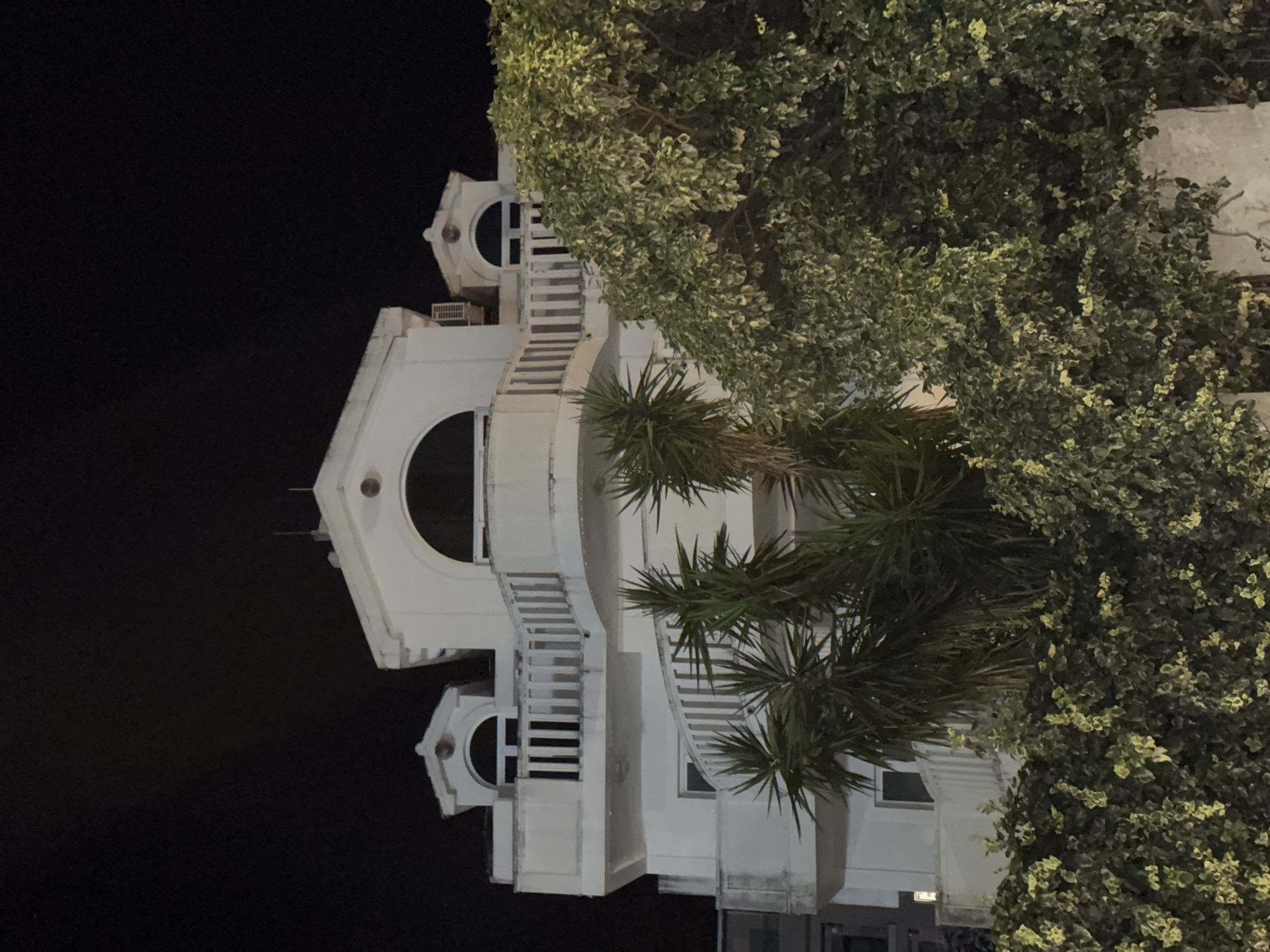The image shows a white, multi-story building at night, with a dark sky as the backdrop. The building features arched windows, balconies with white railings, and a spiral staircase visible on the left side. Lush green foliage, including palm trees and dense bushes, partially obscures the lower levels of the building and a wall in the foreground. An air conditioning unit is mounted on the exterior of the building. The architectural style appears to be Mediterranean or similar, with decorative elements on the roofline. The overall impression is one of a residential or hotel property at dusk or night. While the image is not specifically identifiable as being in Budva, Montenegro, the architectural style and vegetation are consistent with the coastal regions of Montenegro.
