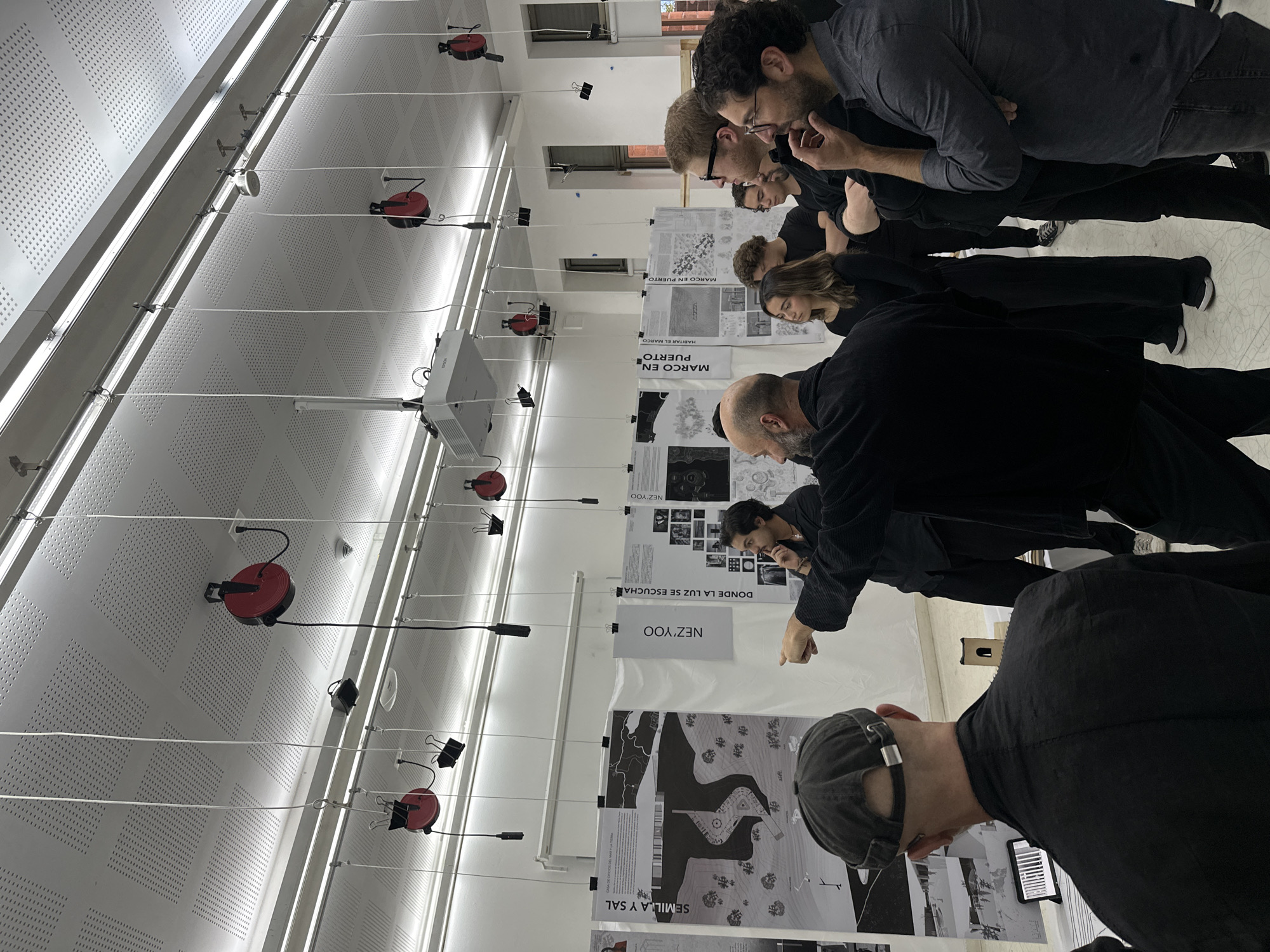 A group of people, mostly men dressed in dark casual attire, are gathered indoors, intently examining a series of architectural or design posters. The setting appears to be a studio, classroom, or exhibition space in Mexico City, Mexico, characterized by a modern ceiling with perforated acoustic panels, linear fluorescent lights, and several red cable reels with white cords suspended from metal rails. A projector is also visible hanging from the ceiling on the left.
The activity centers around the posters, which are hung vertically from the white cords using binder clips. The individuals are leaning in, scrutinizing the details of the presentations. In the foreground, one man in a black cap is seen from behind, while another to his left points towards a poster, suggesting an active discussion or presentation. Other individuals further back are also engaged, looking down at the displayed works. The atmosphere appears focused and collaborative.
Visible text on the posters, in Spanish, includes: "EMILLA Y SAL" (likely "SEMILLA Y SAL," meaning "Seed and Salt"), "NEZ'YOO" (repeated multiple times), "DONDE LA LUZ SE ESCUCHA" ("Where the light is heard"), and "MARCO EN PUERTO" ("Frame in Port" or "Marco in Port"), with parts of "HABITAR EL MARCO" ("Inhabit the frame") also discernable. The posters themselves feature various architectural plans, conceptual drawings, and photographic elements.