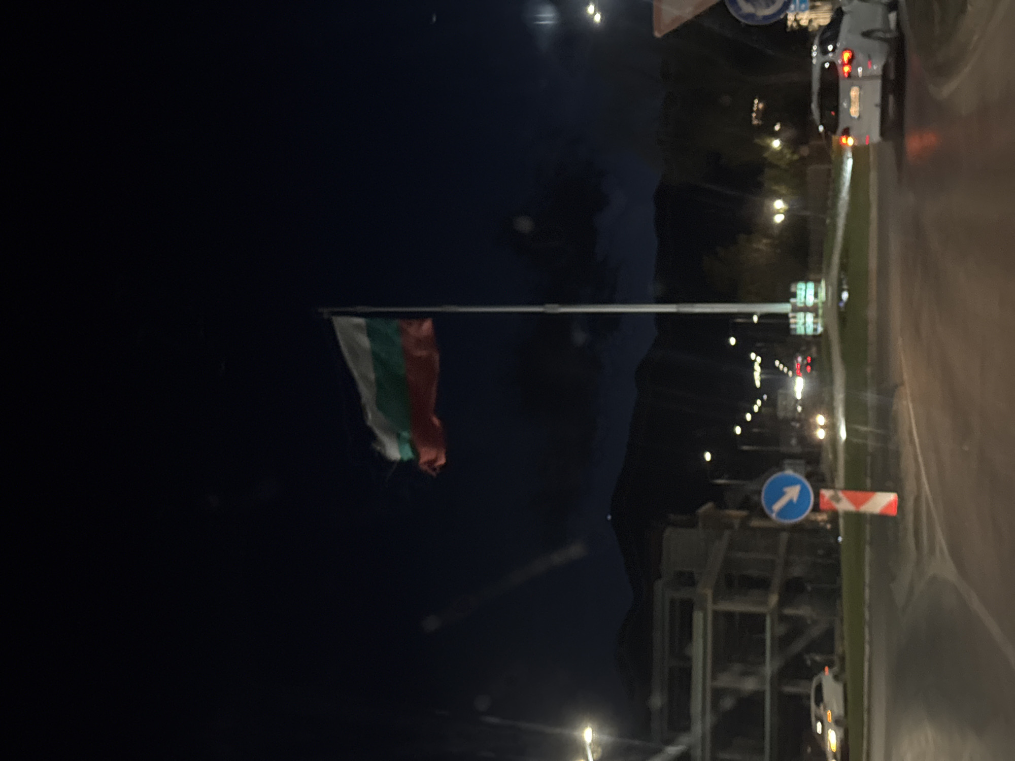 The image shows the Bulgarian flag on a flagpole in Goce Delchev, Bulgaria, illuminated at night. The flag is composed of the colors white, green, and red. The background includes a dark sky, streetlights, road signs (including one with a white arrow on a blue background), and some infrastructure. Vehicles with illuminated tail lights are visible on a road. The photograph appears to be taken at dusk or night.