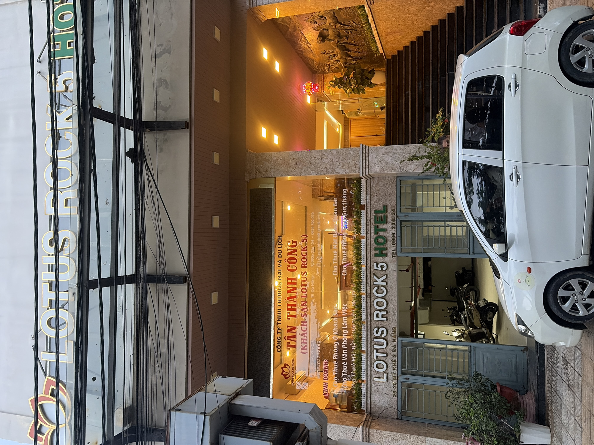 The image shows the exterior of the Lotus Rock 5 Hotel in Da Nang, Vietnam. The hotel's signage is prominently displayed, with the name "LOTUS ROCK 5 HOTEL" in large, illuminated letters. Below the hotel name, the address "23-30 MỸ KHÊ 2 ĐÀ NẴNG" is visible. A white car is parked in front of the hotel's entrance, partially obscuring the ground floor. Motorcycles are parked inside the open entrance. The building has multiple floors with windows and decorative elements. Stairs lead up to an entrance on the right side of the building, illuminated by warm lighting. A mural or relief sculpture is visible on the wall to the right of the stairs. The scene appears to be captured during daylight, with some overhead wires visible in the upper part of the image.
