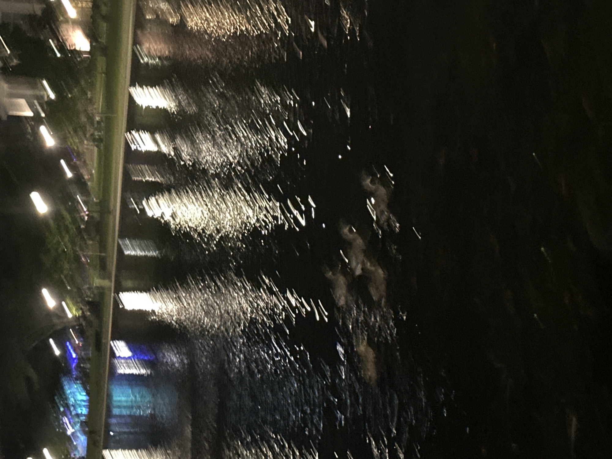 The image depicts a nighttime scene at Big Spring International Park in Huntsville, Alabama. A body of water, likely the Tennessee River, reflects distorted lights from the surrounding urban environment. Buildings and trees are visible in the background, along with streetlights creating elongated streaks of light on the water's surface. The overall impression is one of a vibrant, well-lit city park at night.