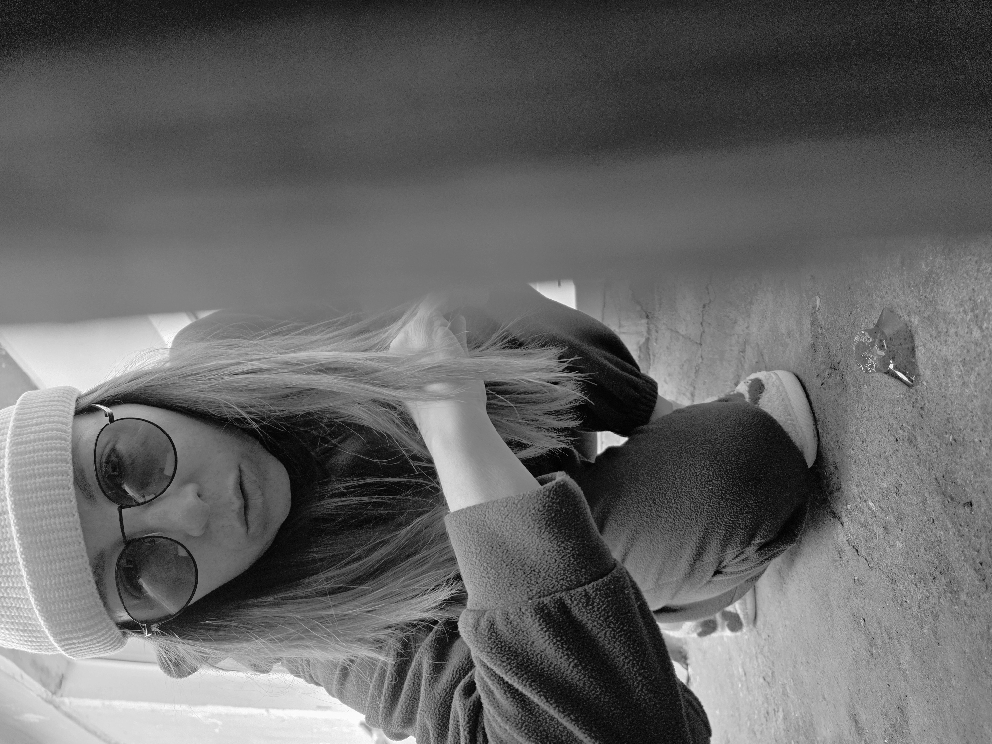 A black and white low-angle shot depicts a person seated outdoors. The person, wearing a light-colored knit beanie and round glasses, looks toward the camera. The person is wearing a dark-colored fleece jacket or sweatshirt. The individual is holding strands of their long, light-colored hair. To the right of the person's legs, resting on what appears to be a concrete surface, is a used candy wrapper. The location is Unknown, Unknown.