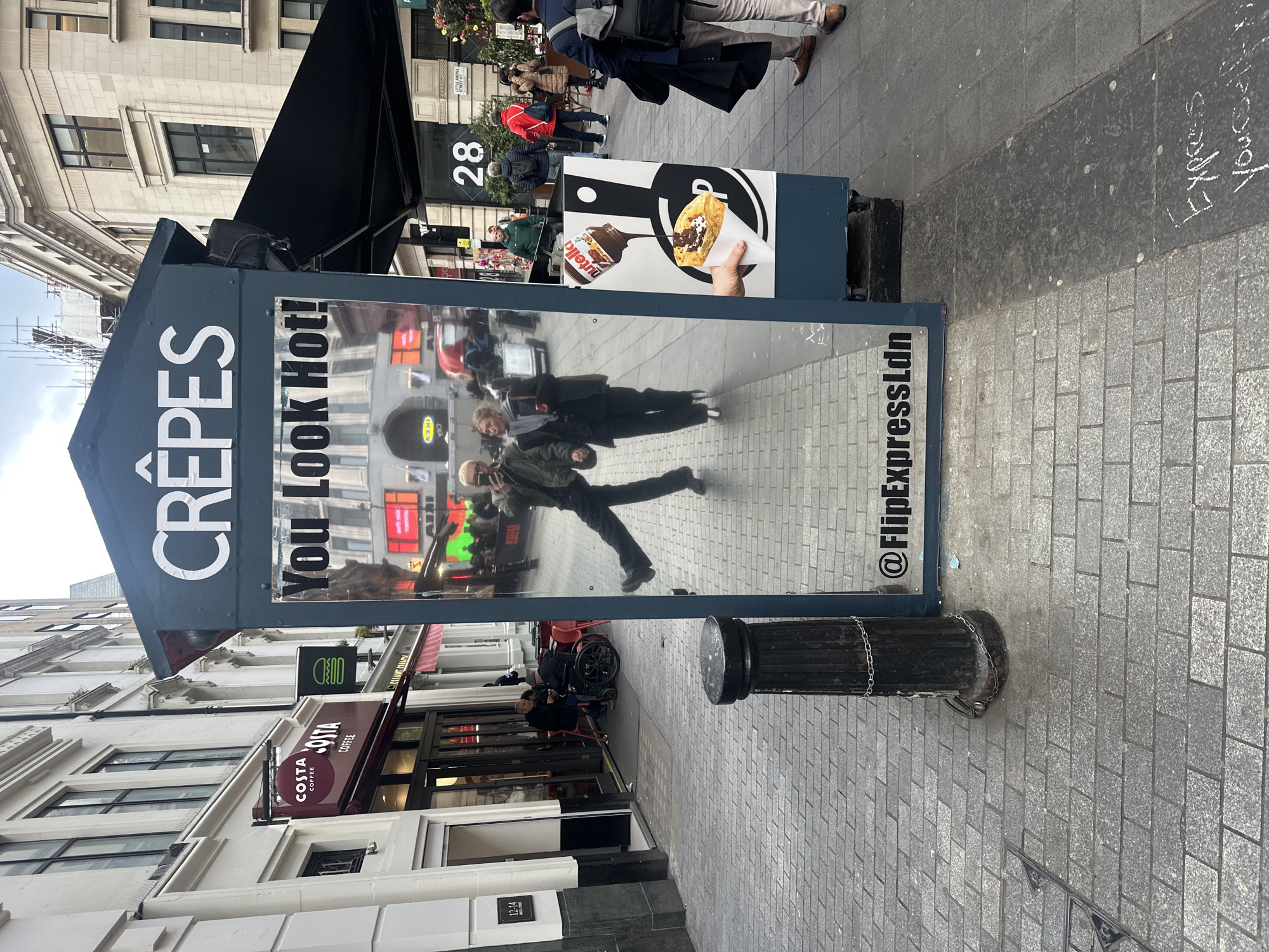 The image captures a street scene in London, United Kingdom. A "CRÊPES" sign with the phrase "You Look Hot!" is prominently displayed. The sign also features a picture of a crêpe with Nutella. The reflection in the sign shows people walking by and the surrounding urban environment. To the left, a Costa Coffee shop is visible. There is a sign next to the crêpes stand with the number "28" on it. A black post with a chain around it stands near the crêpes sign. The text "Express Yourself" is visible on the pavement. The bottom of the sign reads "@FlipExpressLdn".