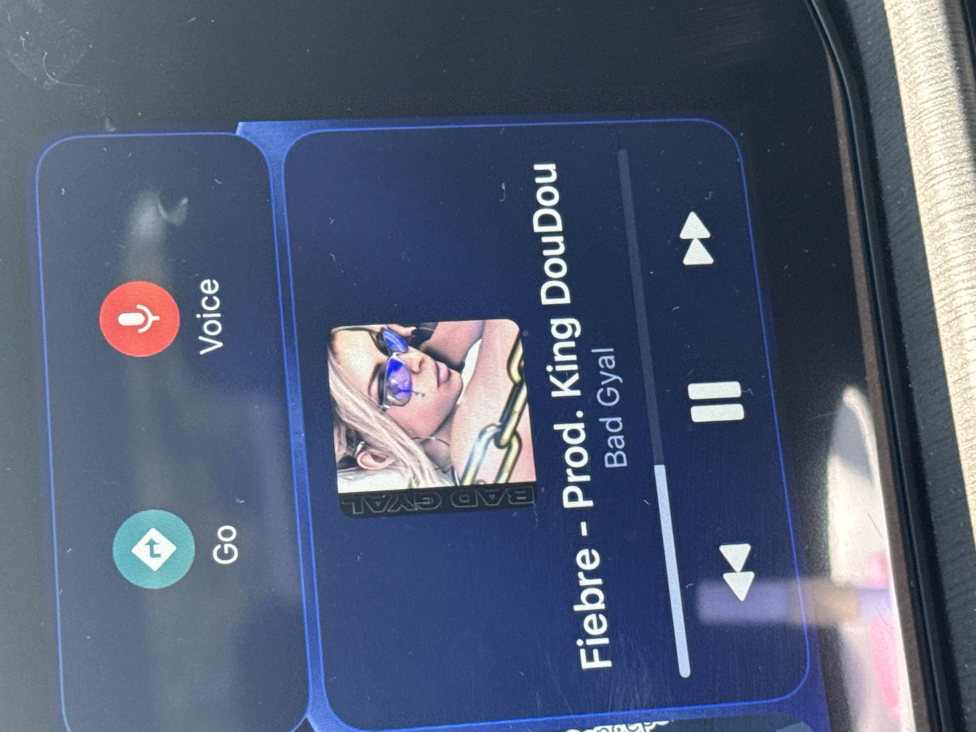 The image shows a smartphone screen displaying the Android Auto interface. Two main sections are visible: one for voice commands ("Voice") and navigation ("Go"), and another displaying music playback information. The music tile shows the song "Fiebre - Prod. King DouDou" by Bad Gyal. The display also shows playback controls like skip forward, play/pause, and skip backward. The photo appears to have been taken indoors, given the presence of a car dashboard. The location, Puebla, Mexico, is not evident from the image content.